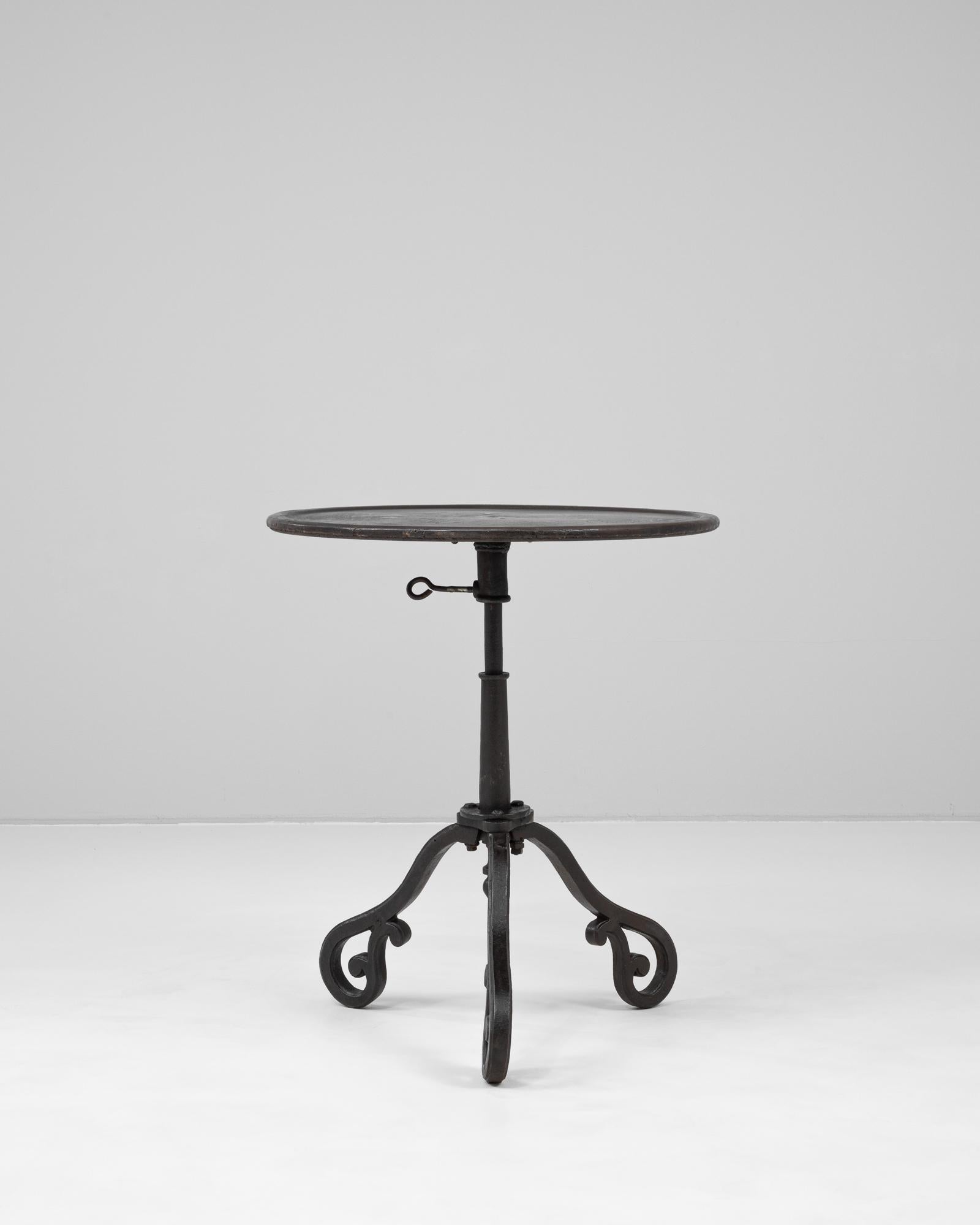 This 1900s French iron side table is a graceful example of industrial design with decorative flair. The round iron top rests on a central adjustable-height column, secured with a classic pin mechanism. Its tripod base features elegantly scrolled