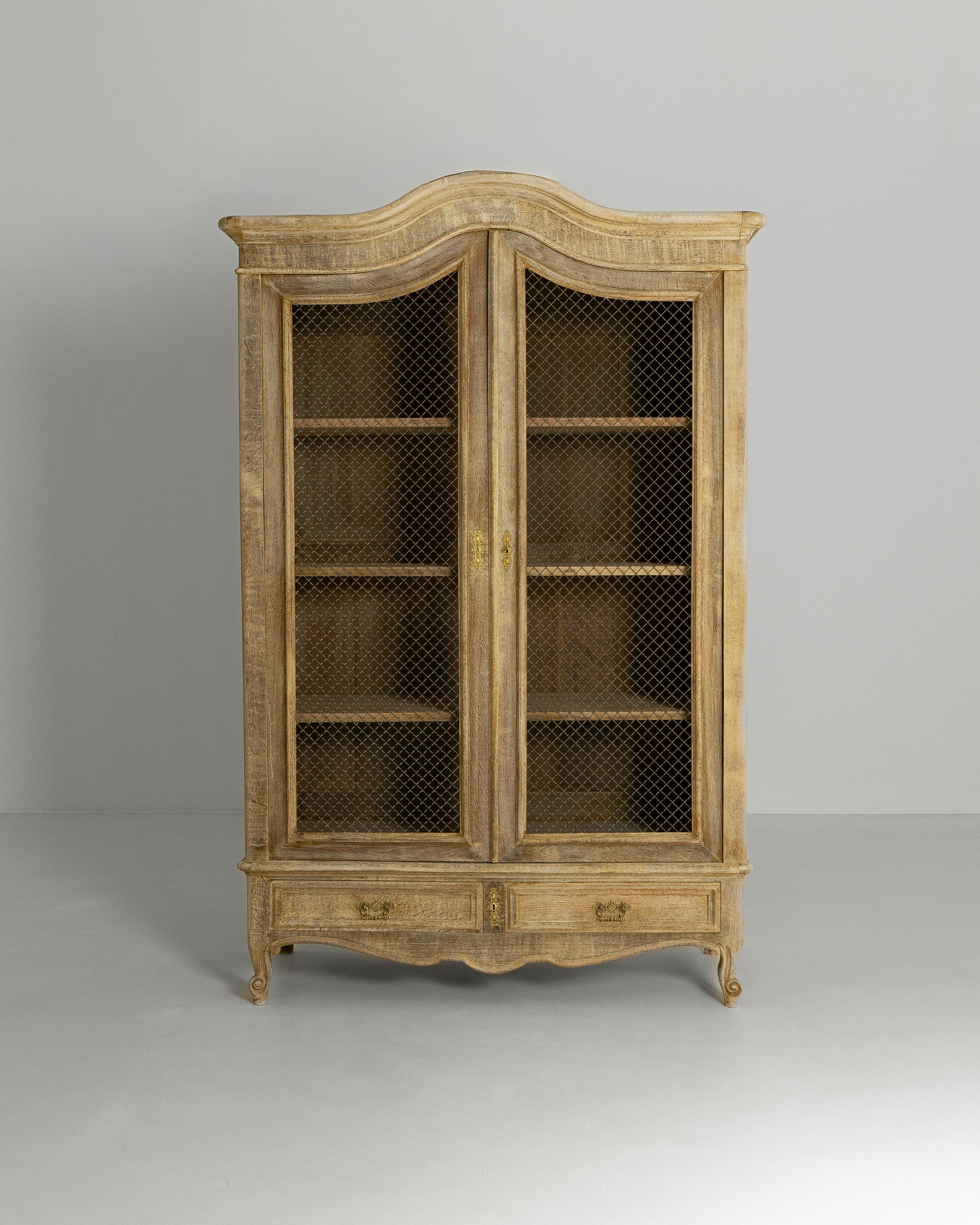 This early 1900s French Louis XV–style oak vitrine exemplifies the graceful curves and refined ornamentation associated with the period. Crafted from solid oak, the cabinet features a softly arched cornice, flowing lines, and cabriole legs that lend