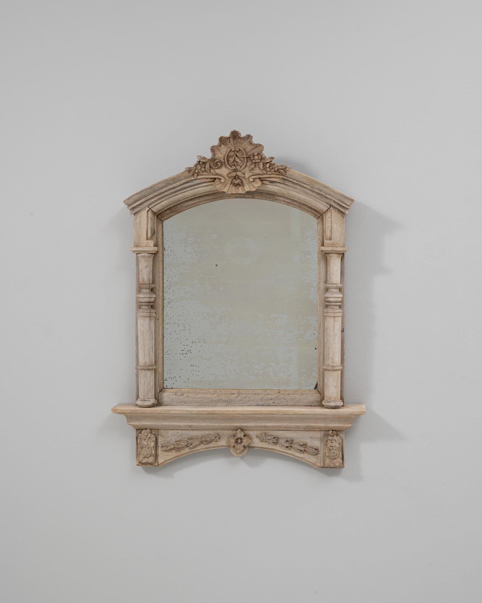 This 1900s French Louis XVI carved oak mirror with shelf features an elegant architectural silhouette and refined decorative details characteristic of the period. The arched frame is topped with an intricately carved crest of scrolls and foliage,