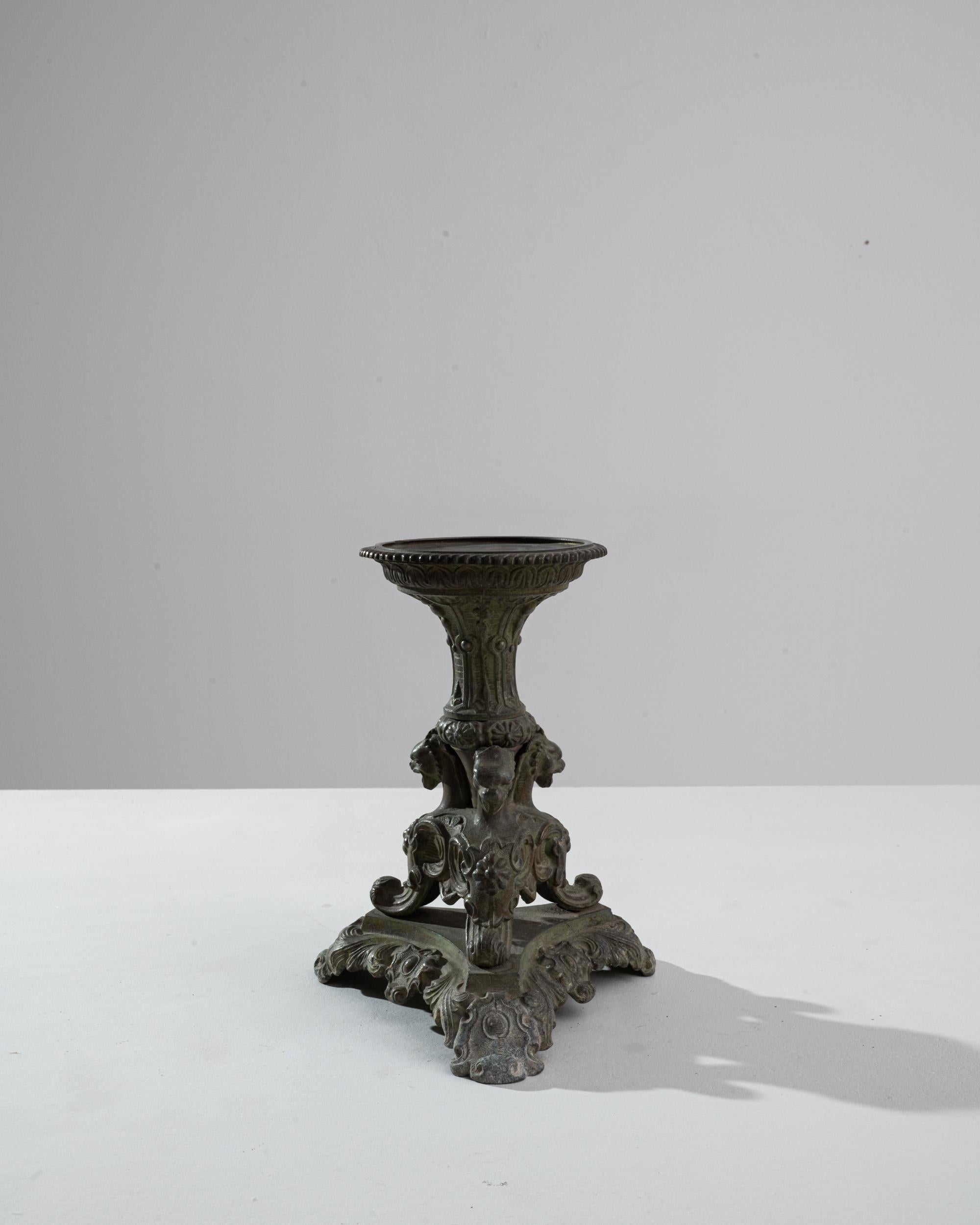 A metal candlestick created in 1900s France. This lavishly detailed candlestick made from cast iron emits a solemn and engaging aura. Mysterious creatures, painstakingly detailed, emerge from each end of its tripod of legs, reinforcing the neo