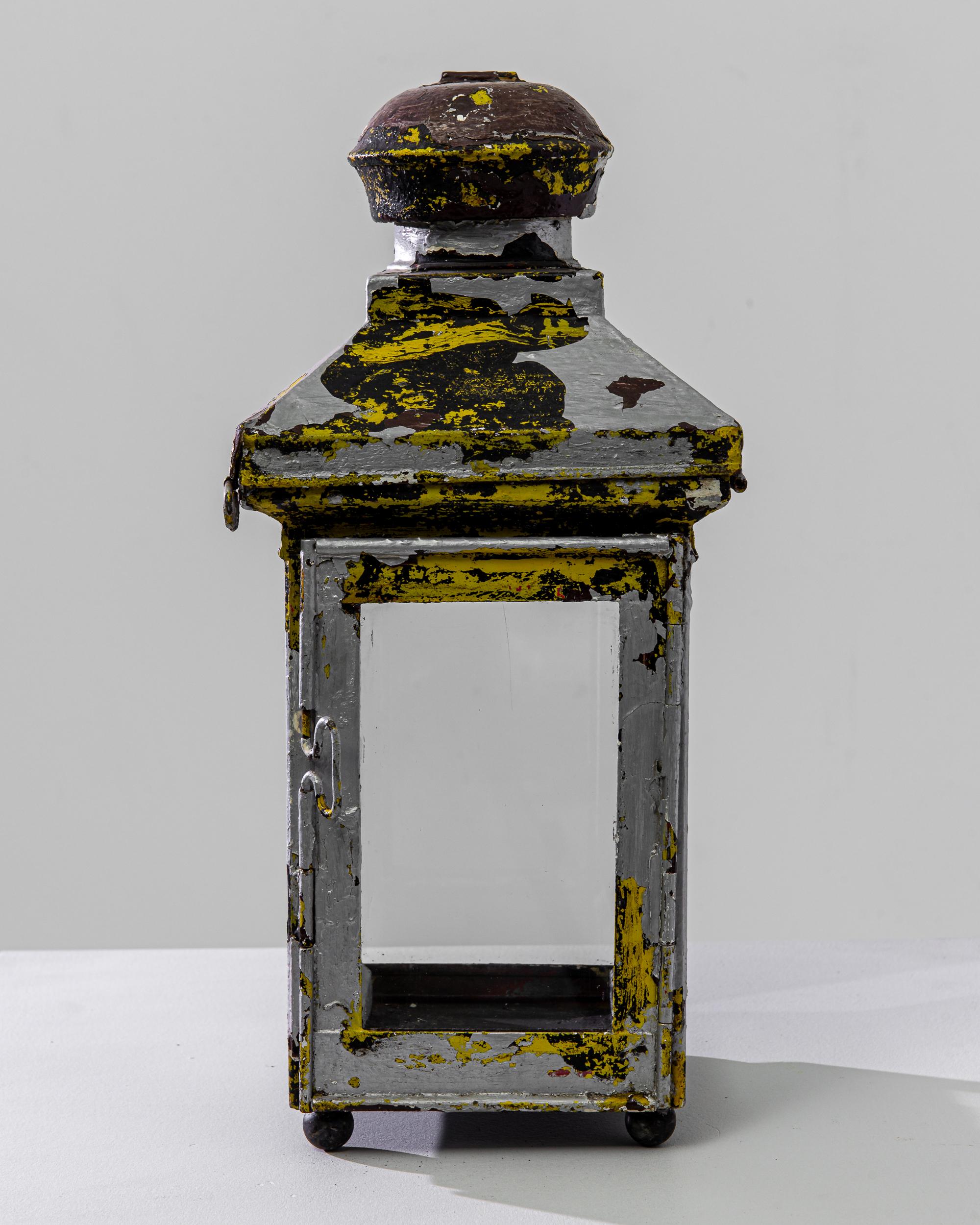 Behold a genuine artifact of the 1900s, a French metal lantern that exudes the charm and character of a bygone era. This piece stands as a testament to history, its patina a narrative of its past, with layers of chipped yellow and gray paint that