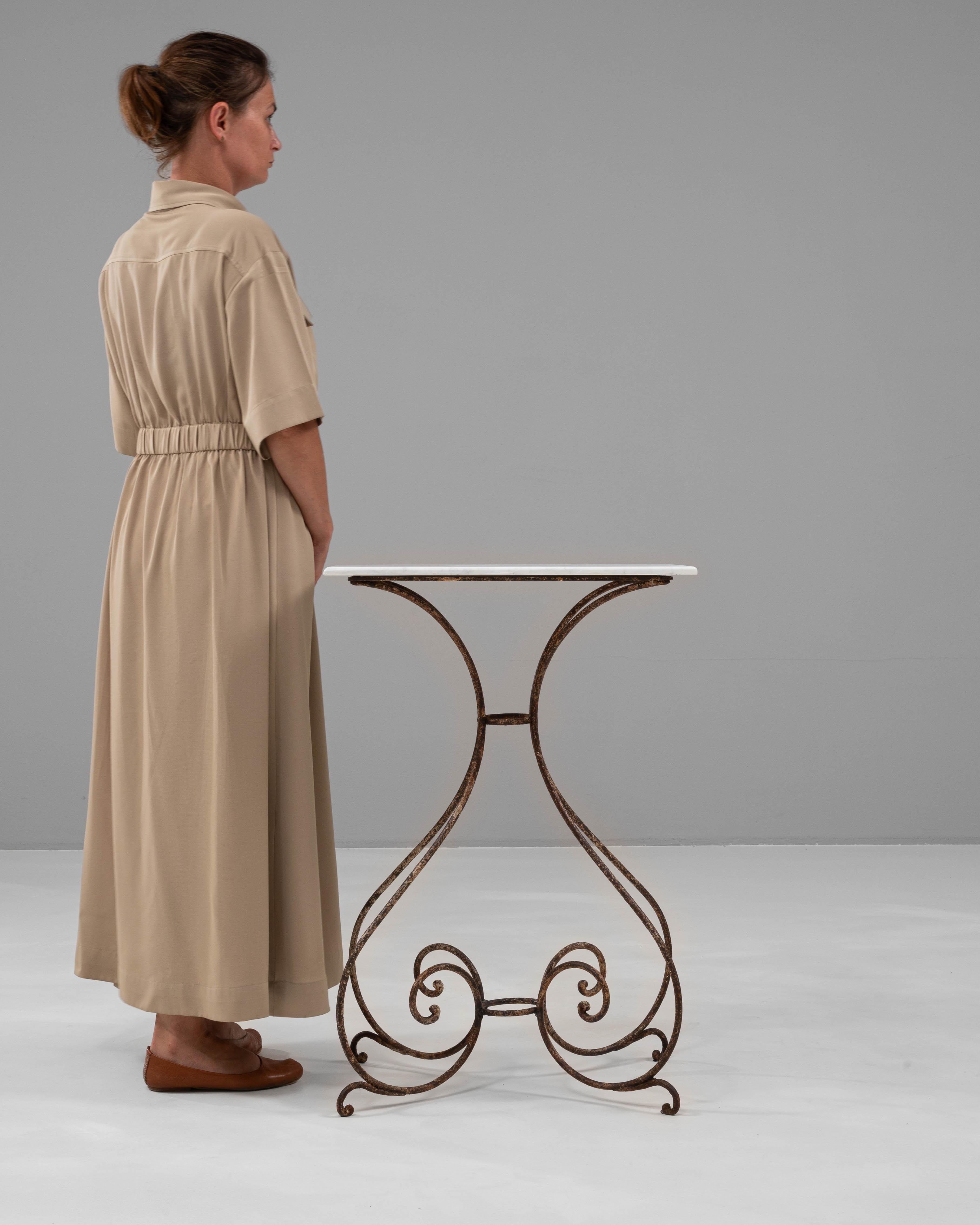This 1900s French metal table with a marble top is a stunning example of timeless craftsmanship. The ornate wrought iron base features intricate scrollwork, lending the piece an air of sophistication and elegance. Its weathered patina showcases its