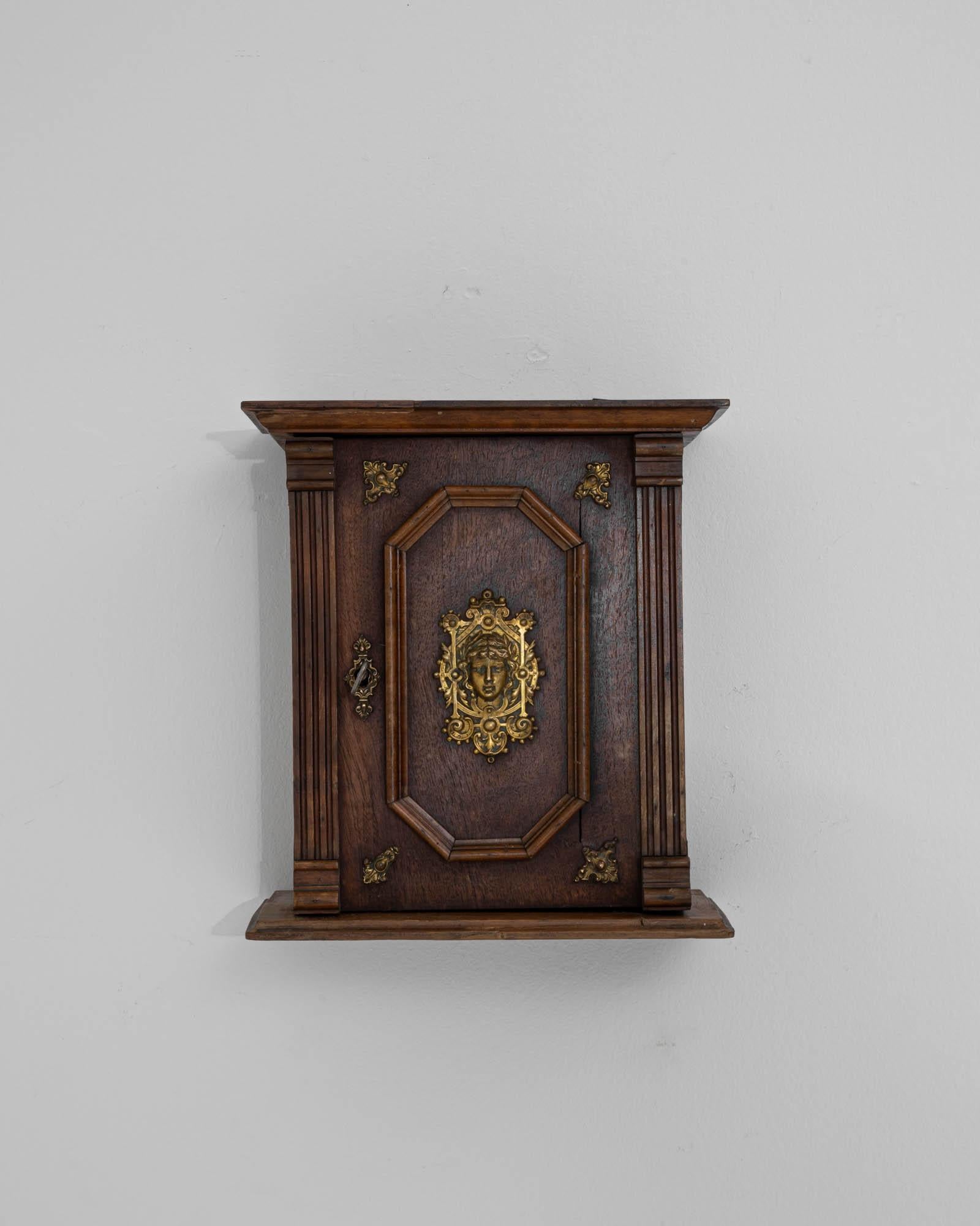 A wooden miniature wall cabinet created in France circa 1900. The golden visage that has been cast and affixed to the center of the panel door of this cabinet meets one with a stoic and calming gaze. The spiraling details floral motifs punctuated in