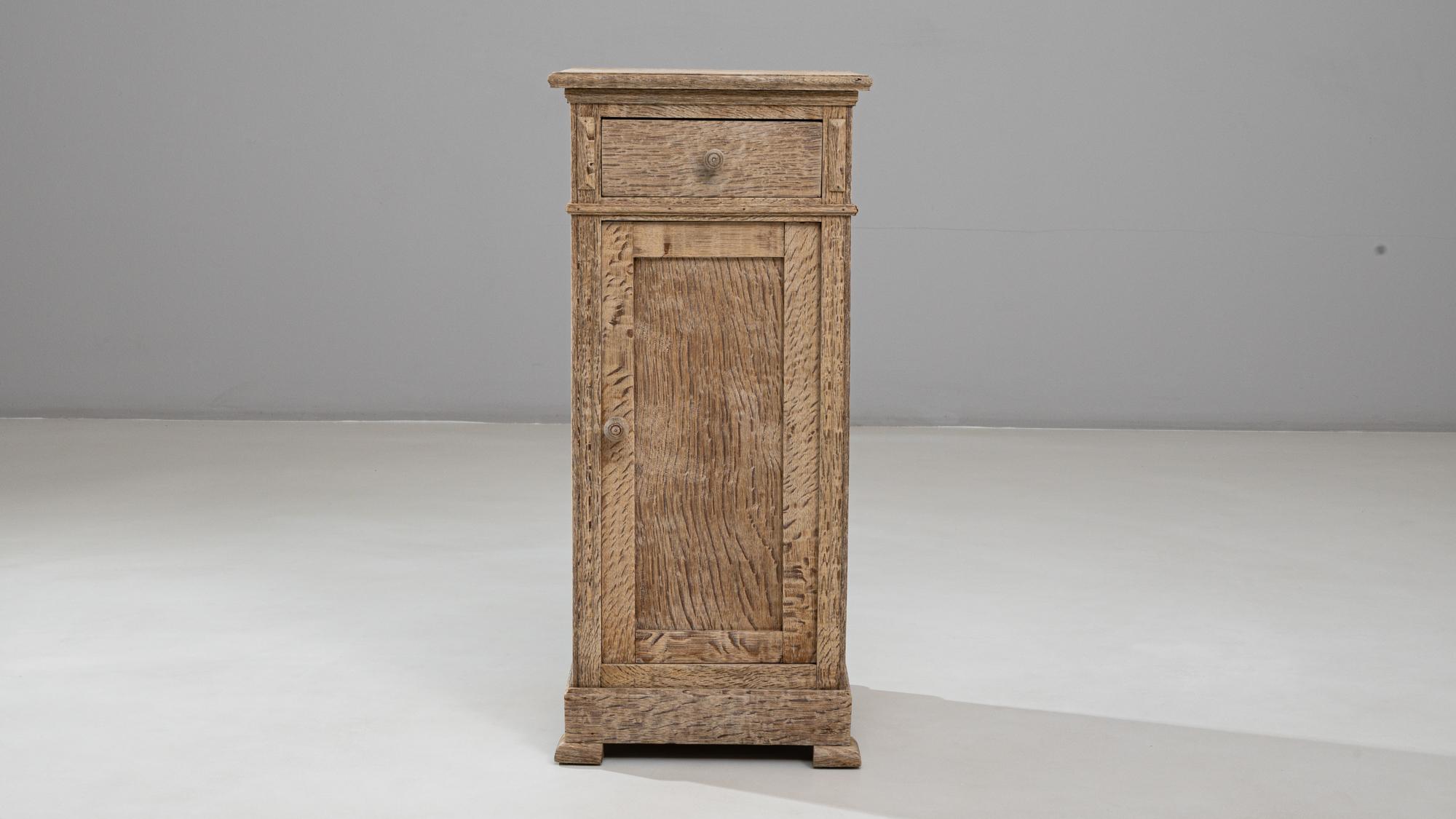 This 1900s French oak bedside table presents a simple, rustic elegance rooted in traditional craftsmanship. Built from solid oak with a warm, weathered patina, it features a single upper drawer with a round wooden pull and a lower cabinet door