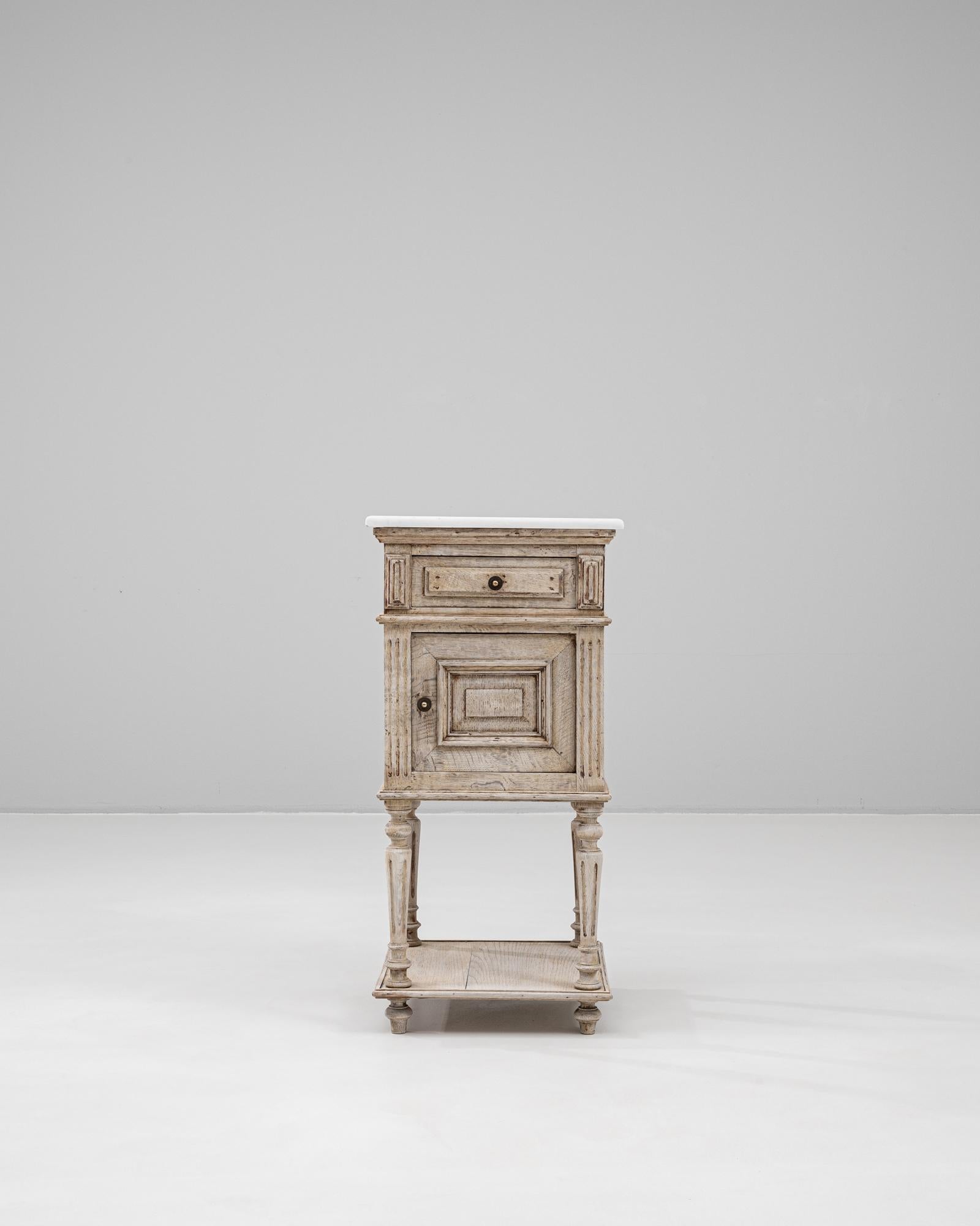 Crafted in France at the turn of the 20th century, this oak bedside table features a timeless silhouette enriched with refined neoclassical details. The light oak frame is gently patinated, highlighting the wood’s natural grain, while a white marble