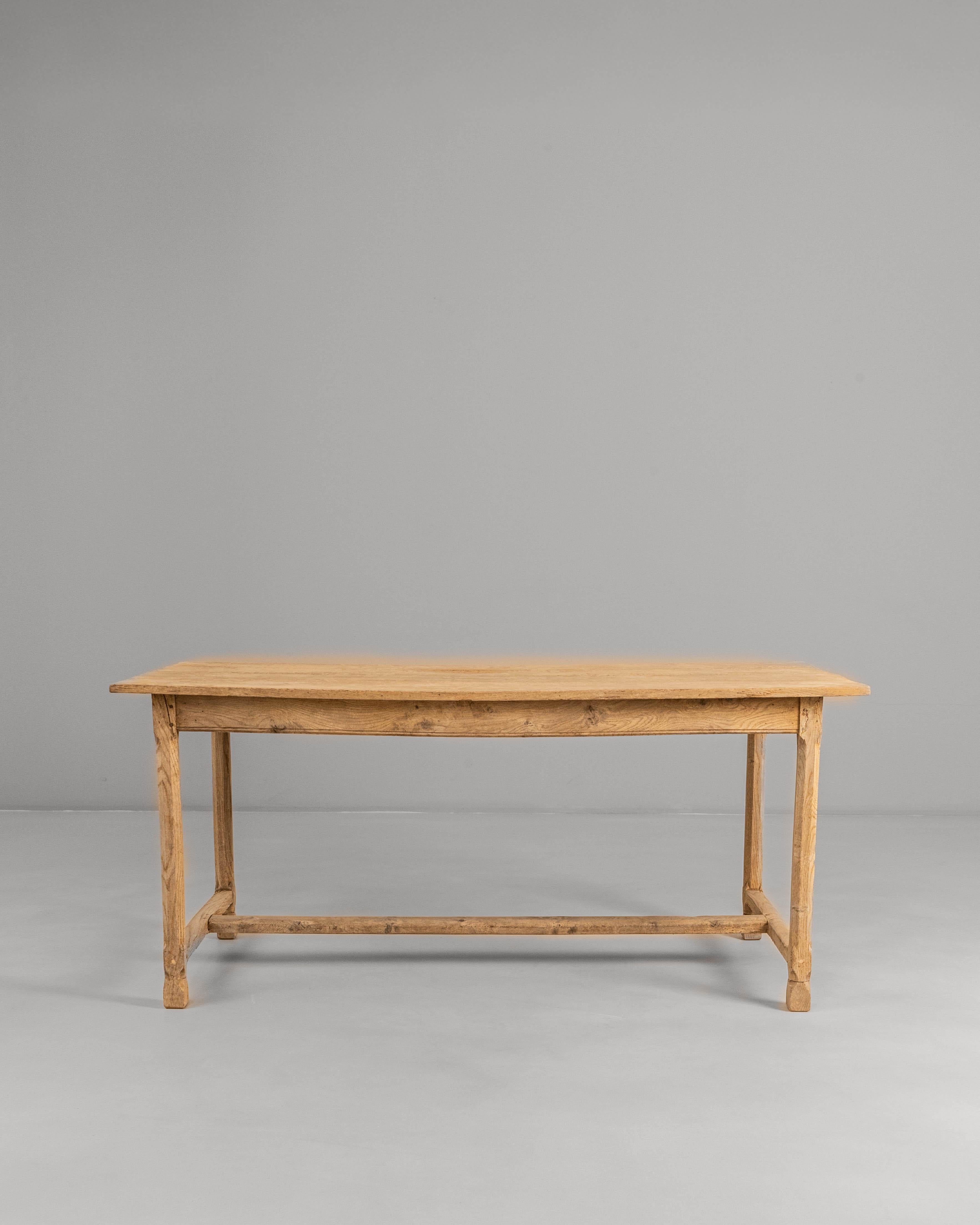 1900s French Oak Dining Table. This understated early 20th-century dining table is crafted from solid oak, showcasing a warm, natural patina that highlights the grain and subtle irregularities of the wood. Its long rectangular top rests on tapered
