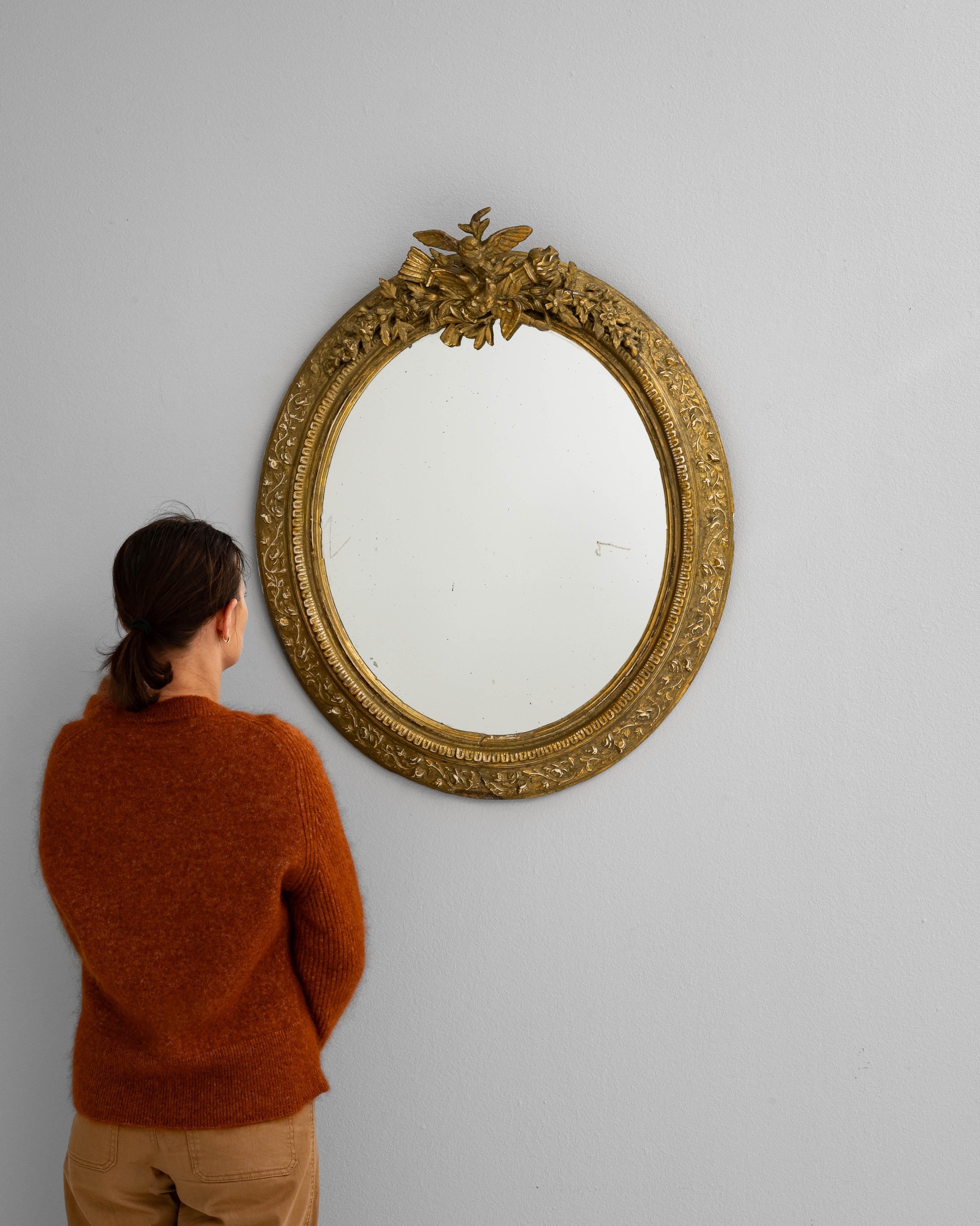 Ce miroir ovale français des années 1900 est un exemple élégant de l'artisanat du début du siècle, avec un cadre en bois sculpté orné de gesso doré. Le cadre est orné de motifs floraux et feuillus le long du périmètre, culminant dans un écusson orné