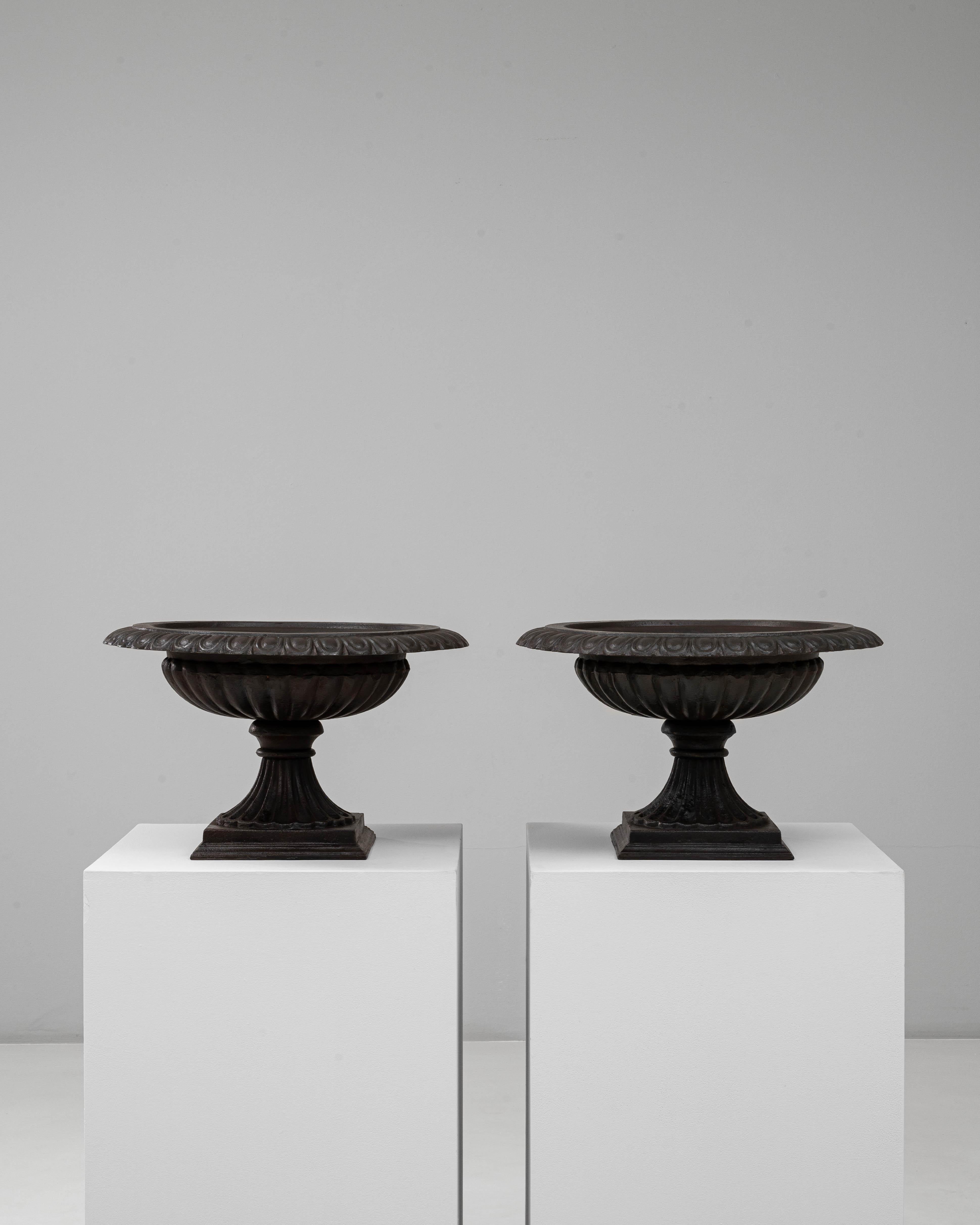 Questa coppia di fioriere a urna in ghisa del 1900 incarna la raffinatezza neoclassica e l'eleganza senza tempo del giardino. Ogni urna presenta una ciotola scanalata e poco profonda con un ampio bordo decorativo ornato da motivi di foglie