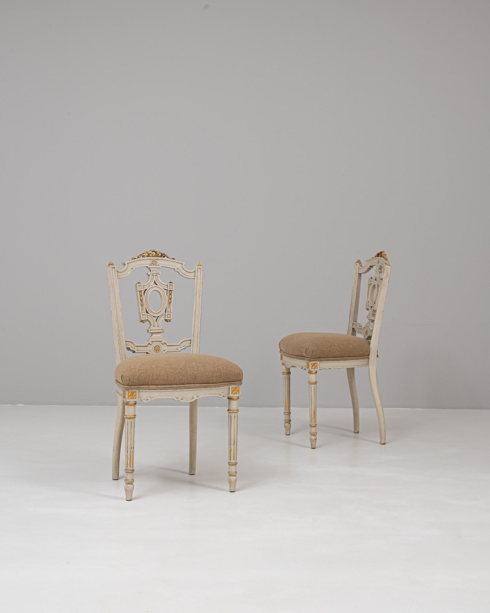 Questa coppia di sedie dorate Luigi XVI patinate bianche del 1900 cattura l'eleganza neoclassica dello stile Luigi XVI con dettagli raffinati e forme aggraziate. Ogni sedia è caratterizzata da una struttura in legno intagliato con una finitura