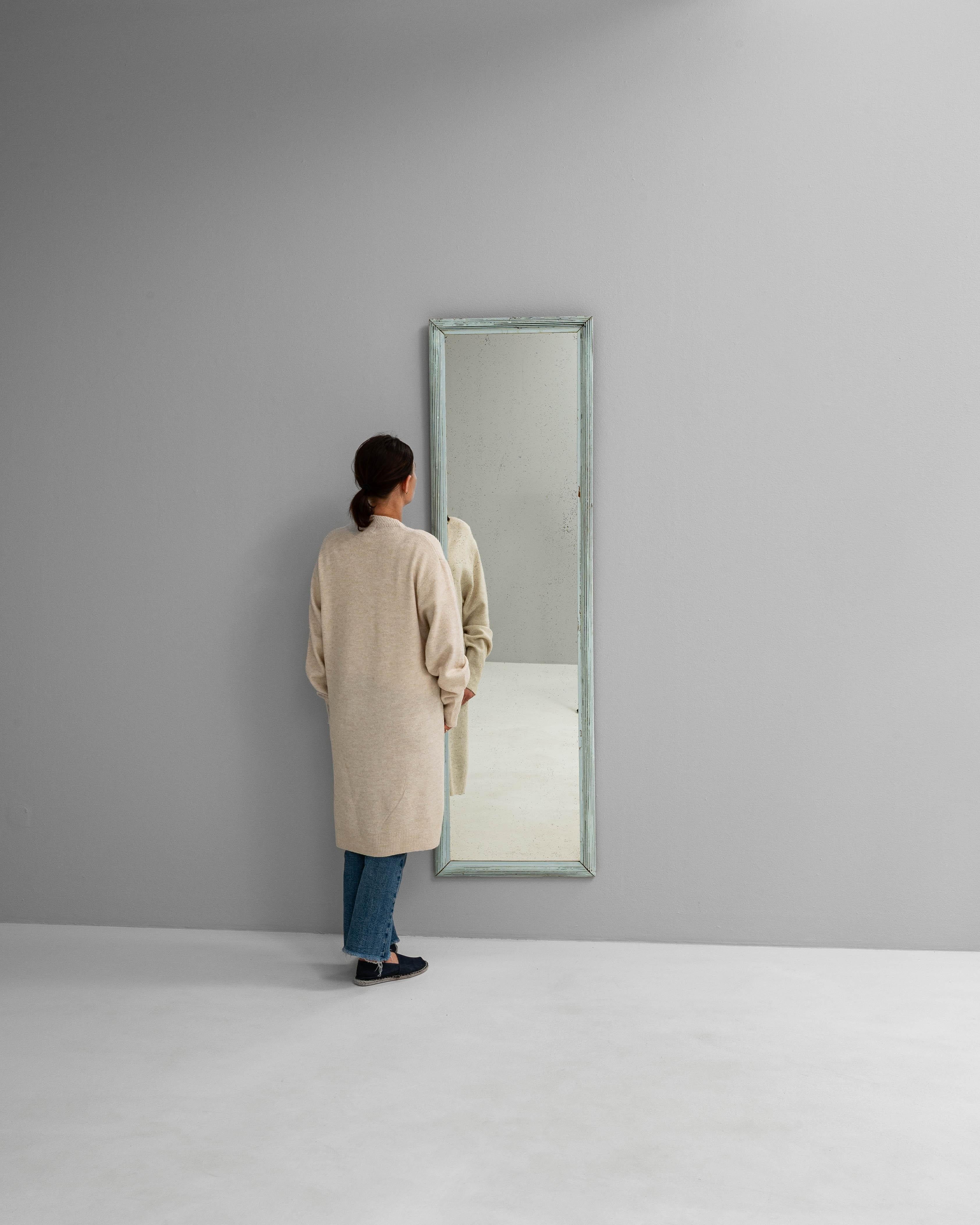 This 1900s French floor mirror features a slender vertical form with a subtly beveled wooden frame finished in a soft, weathered patina of pale blue and white tones. The aged paint reveals hints of the natural wood beneath, lending a charming,