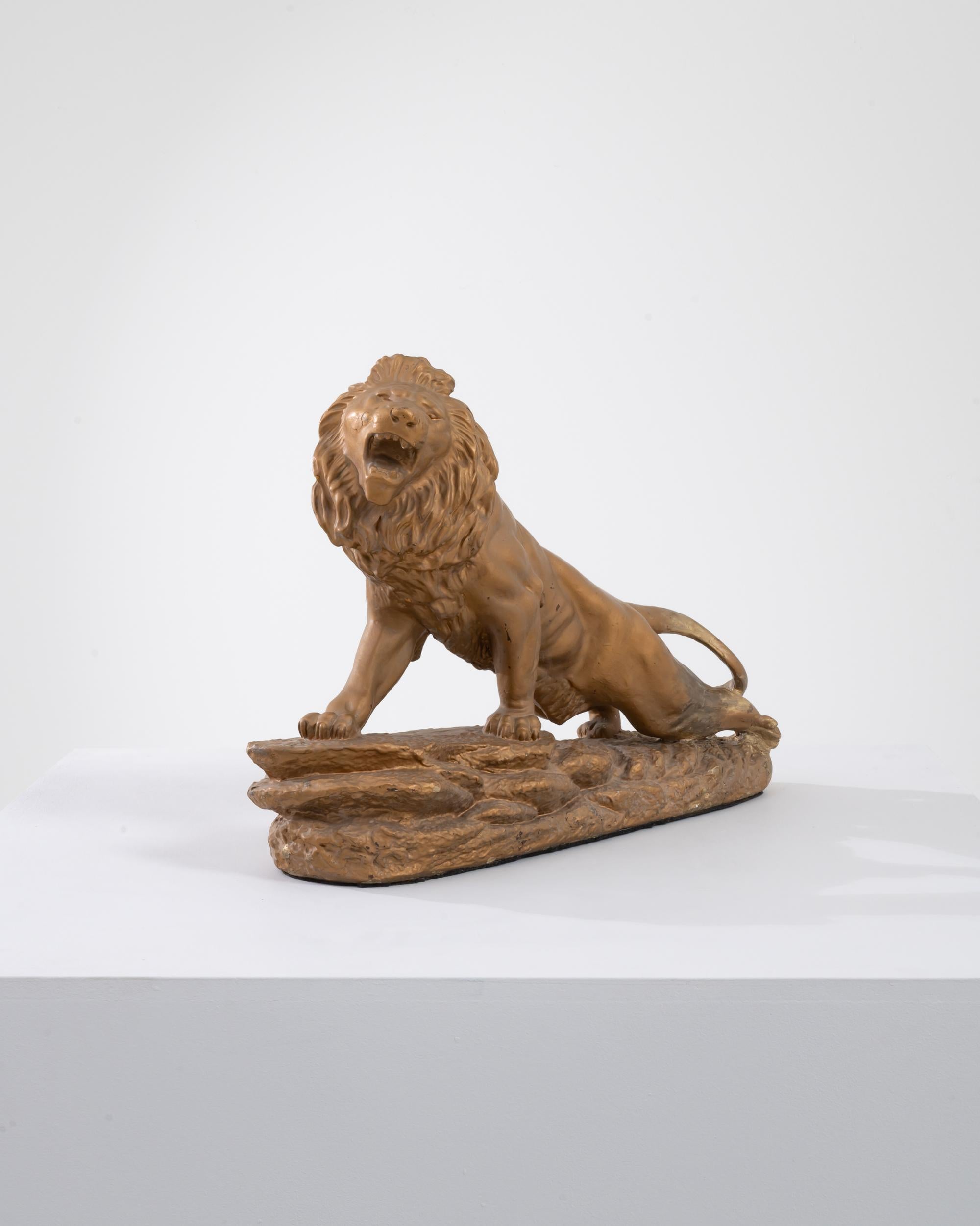 A painted plaster sculpture from 1900s France. This sculpture depicts a lion climbing up to a cliff's edge, with legs stretched back releasing a ferocious roar. The gold paint that coats the plaster cast sculpture has chipped here and there, adding