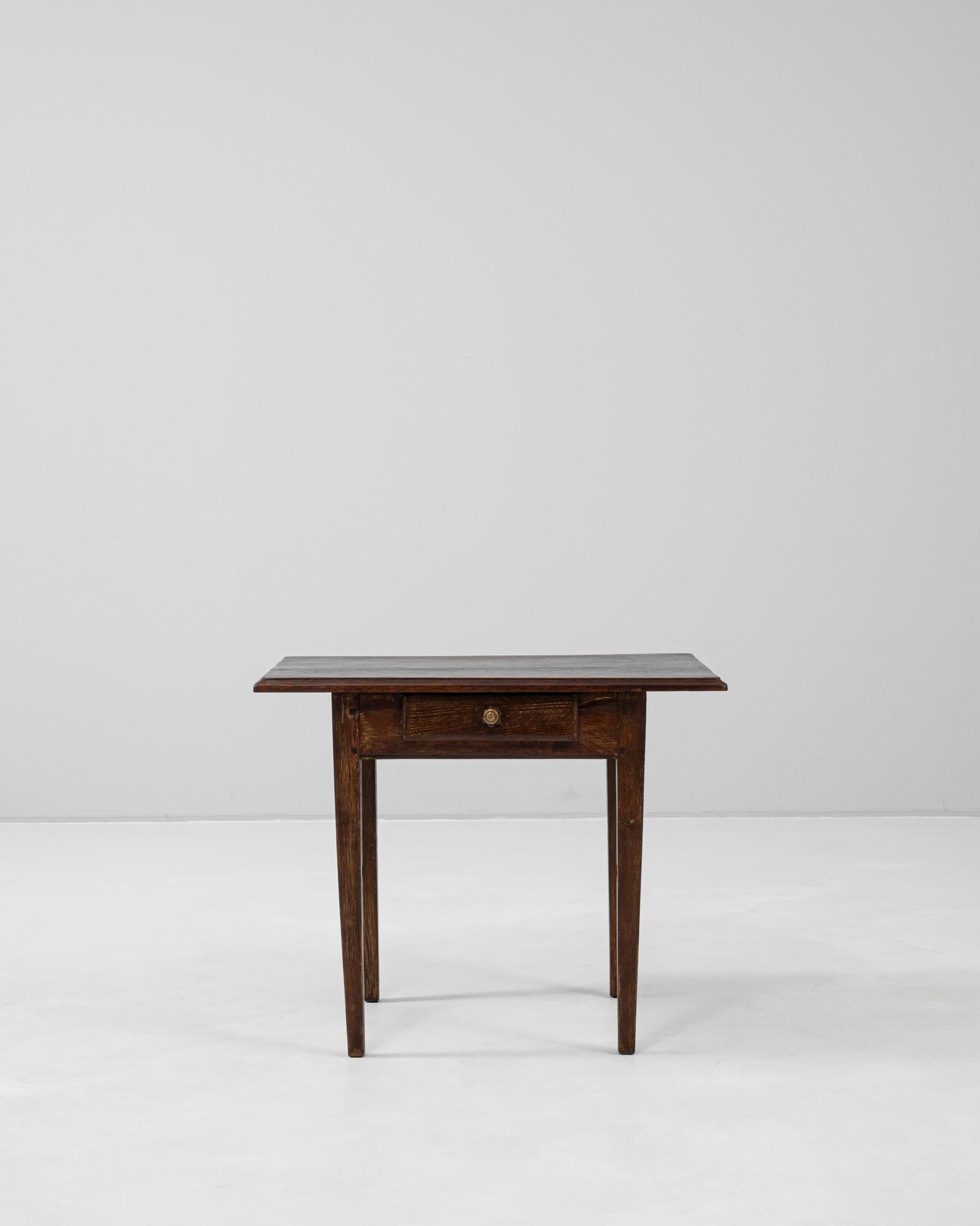 This early 20th-century French side table features a simple, timeless silhouette with tapered legs and a single drawer for added functionality. The rich, dark finish highlights the natural grain and character of the wood, while the drawer’s brass