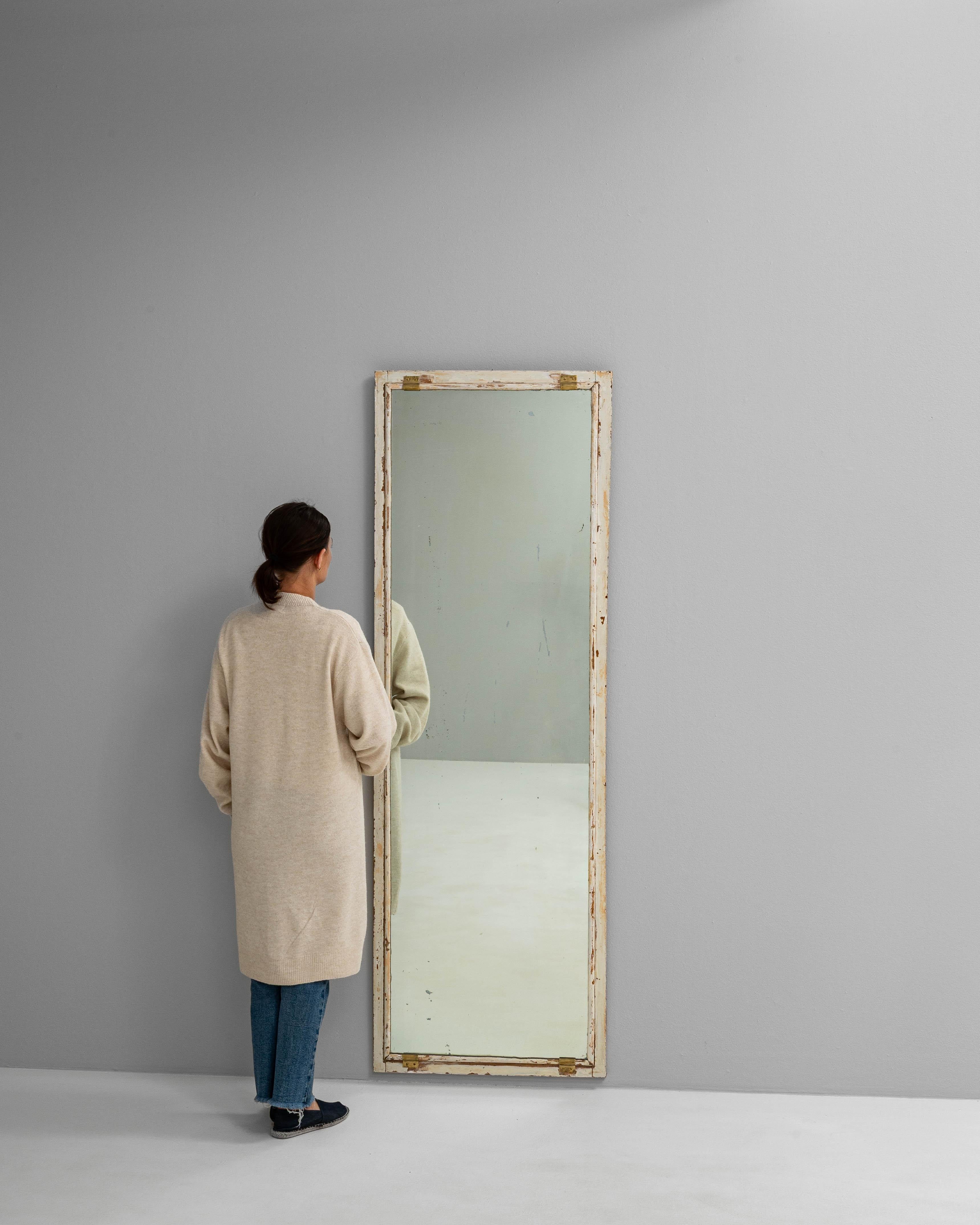 This 1900s French floor mirror features a tall, slender profile framed in wood with a beautifully weathered white patinated finish. The frame shows signs of natural age and wear, with areas of exposed wood and remnants of old paint adding authentic