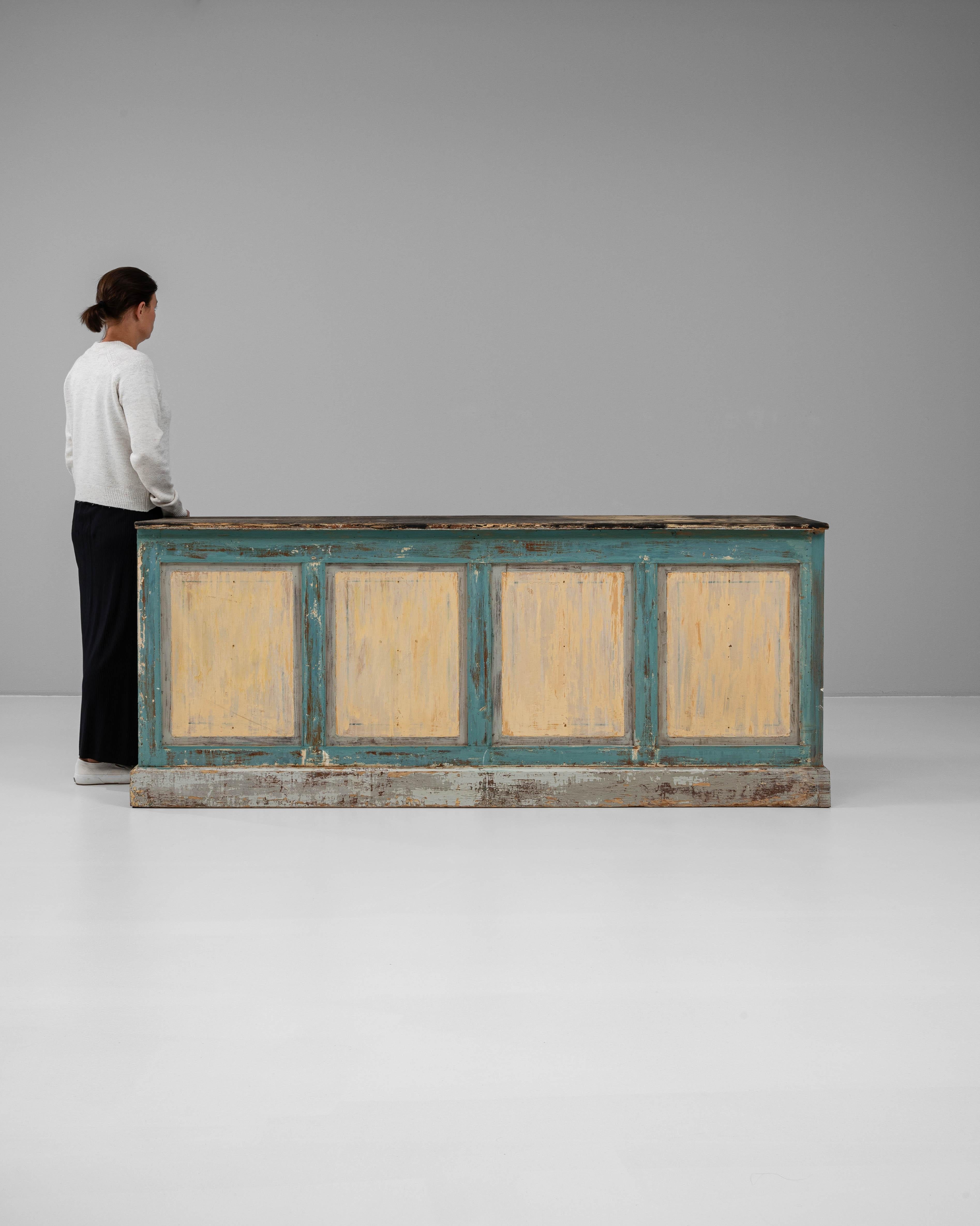 This 1900s French wooden bar is a richly patinated statement piece, brimming with character and old-world utility. The front showcases four framed panels in a two-tone painted finish—muted ochre framed by weathered teal—that has naturally worn over