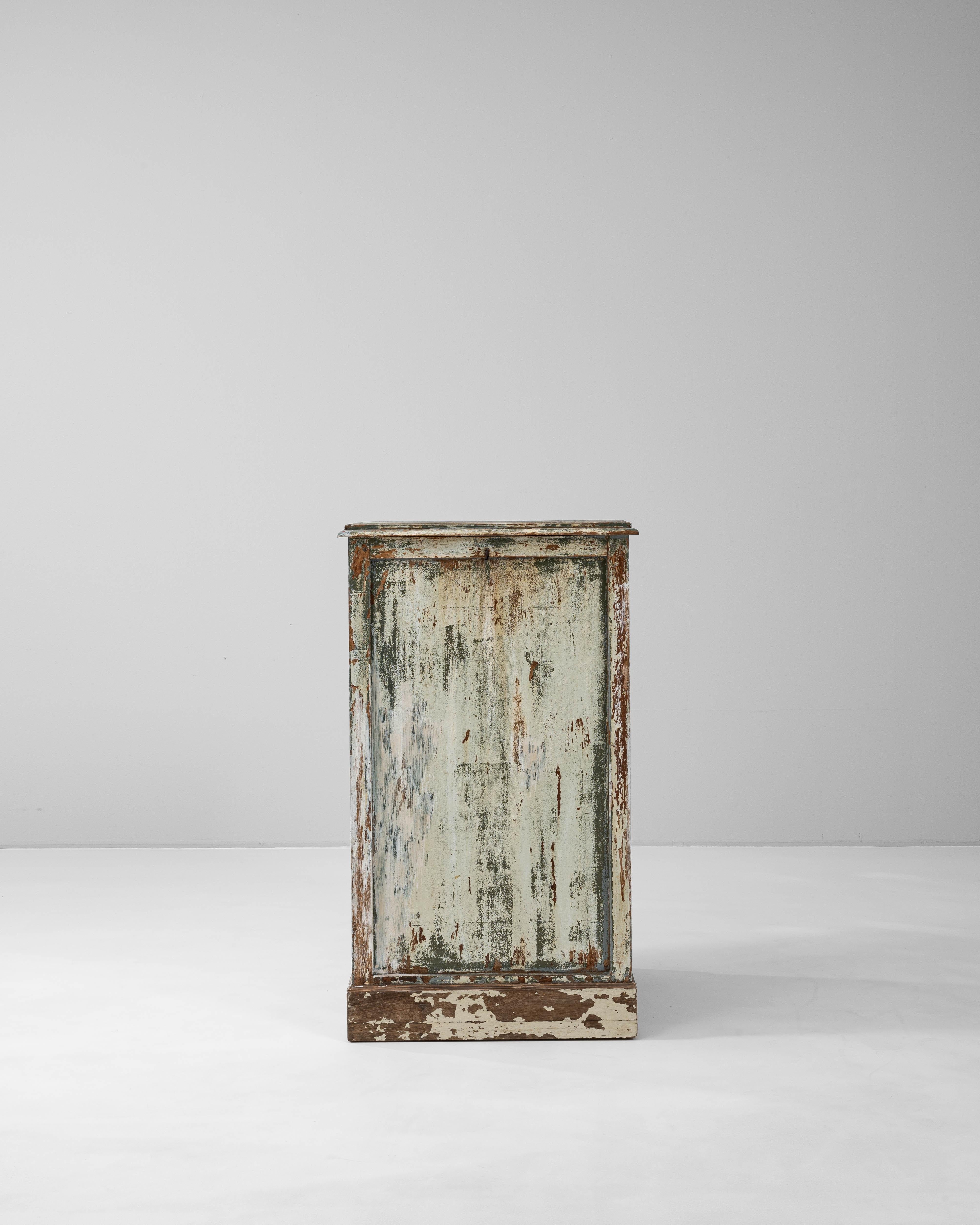 This 1900s French wooden chest is a striking example of timeworn elegance, featuring a richly patinated surface in layers of pale green, white, and natural wood tones. Its tall, rectangular form is enhanced by recessed paneling and a subtly molded