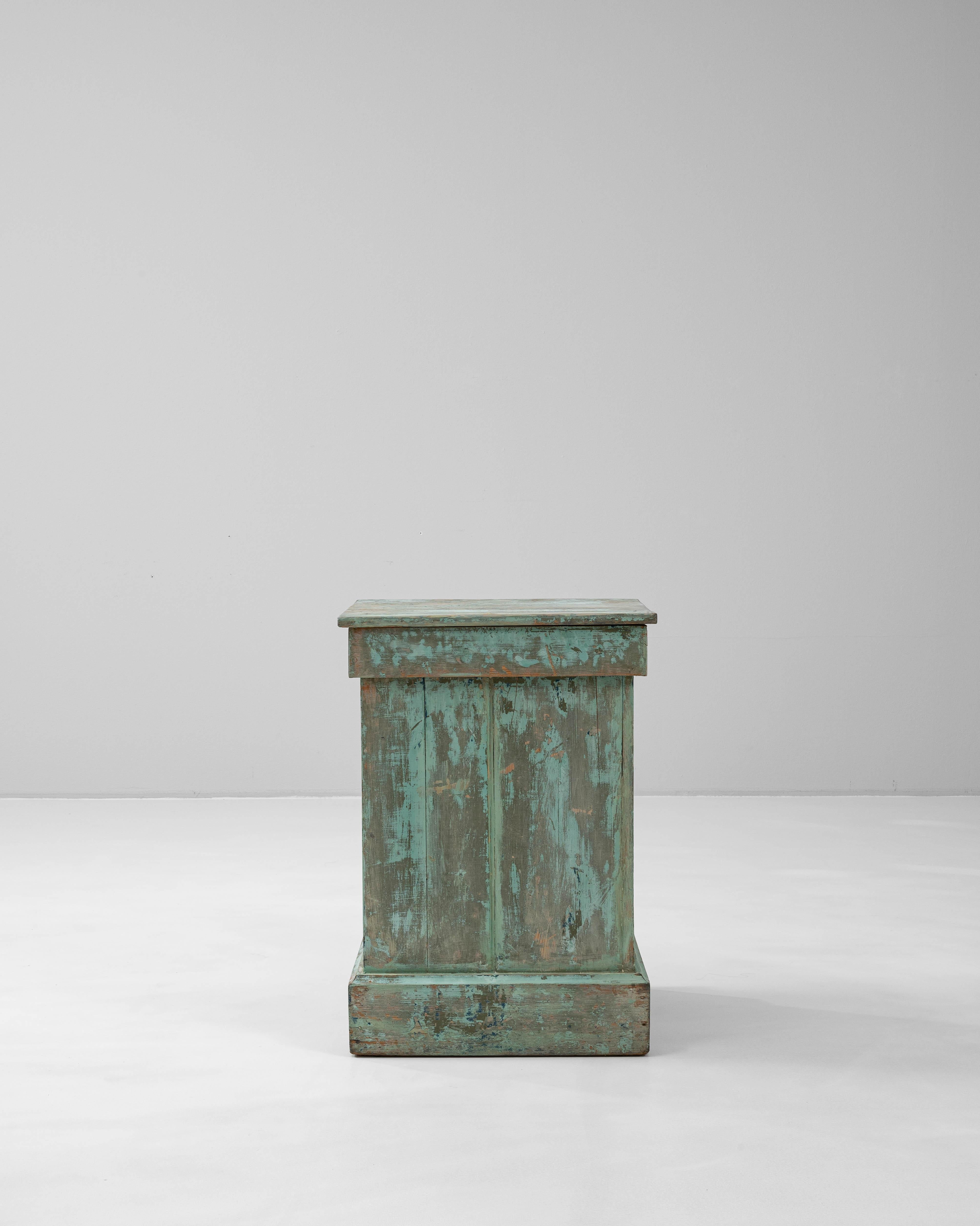 This 1900s French wooden pedestal features a beautifully worn patina in soft turquoise and earthy tones, evoking a rustic charm that speaks to its age and history. Crafted from solid wood, the piece has a simple rectangular form with subtle molding