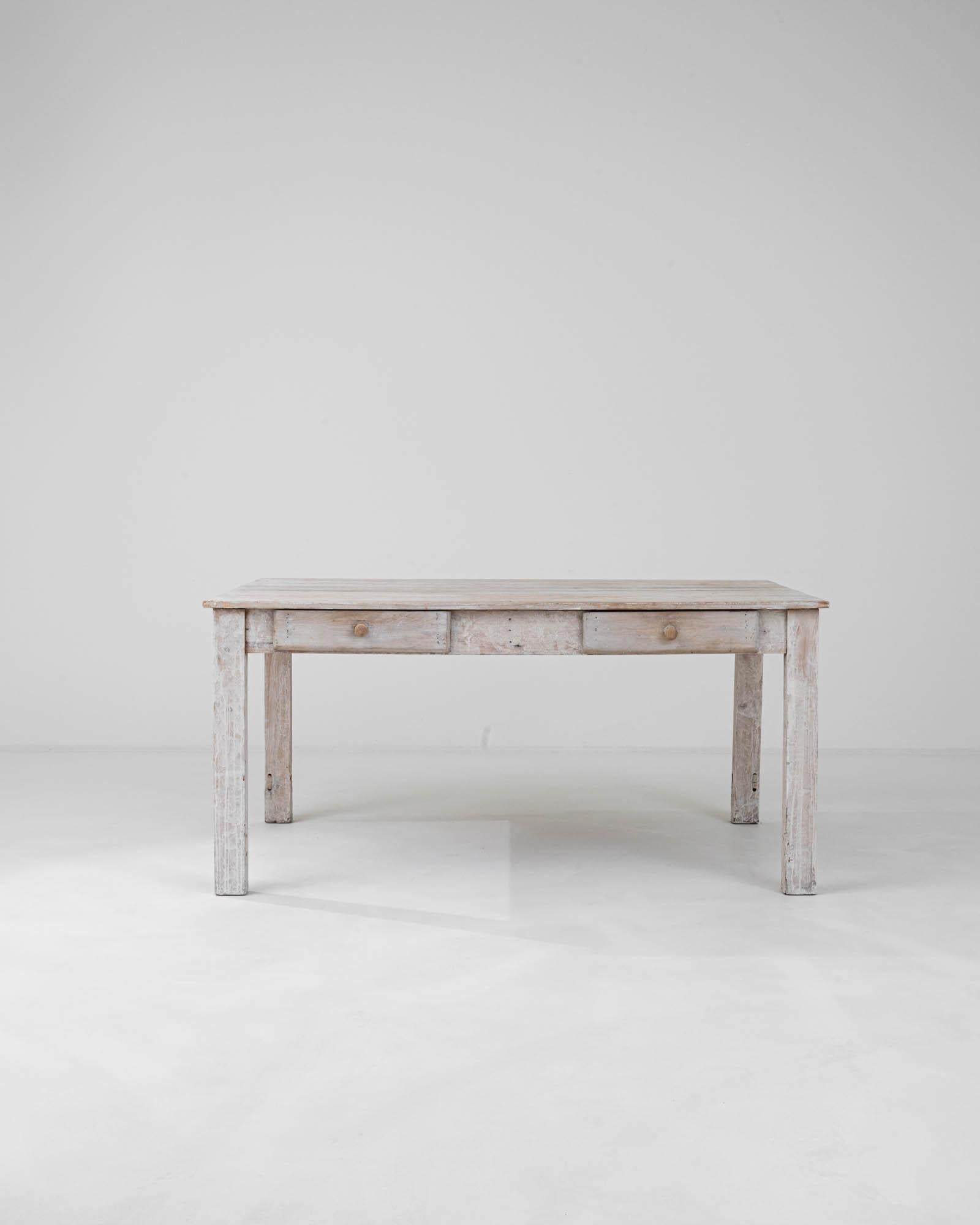 This 1900s French wood table, bearing the graceful wear of white patination, tells the story of a bygone era. The beautifully aged surface, with its natural wood grain peeking through the distressed finish, invites a sense of rustic charm into any