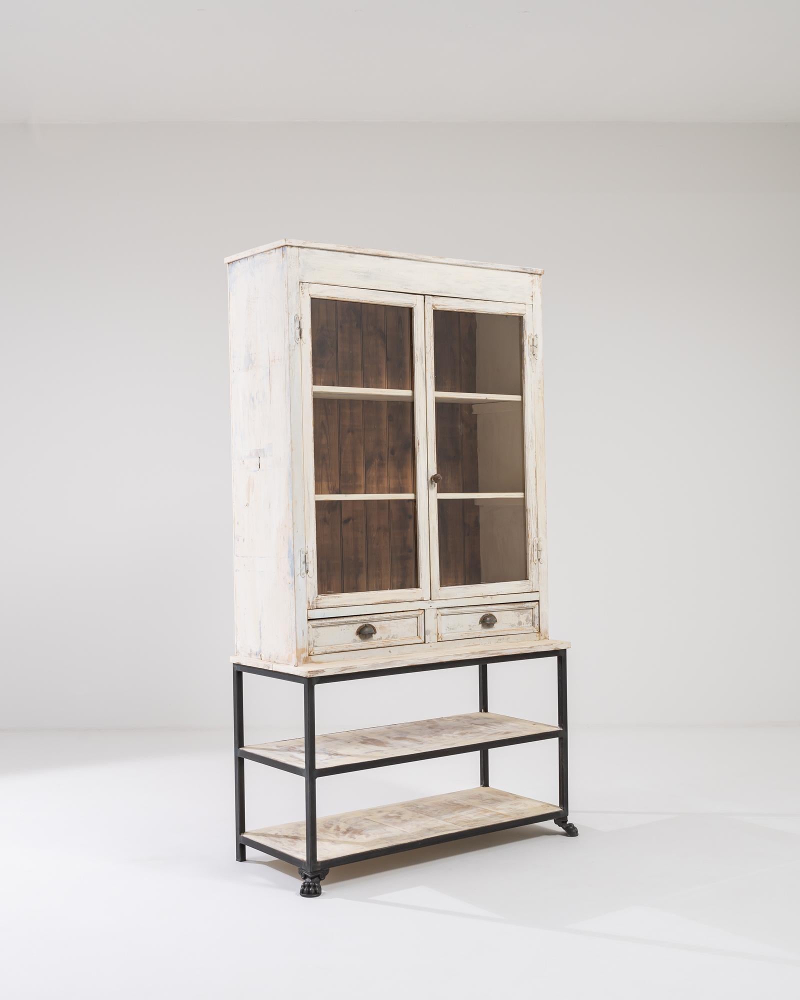 A wooden and metal vitrine from France, circa 1900. This vitrine is composed of two lower shelves, two drawers and two shelves which rest inside glass paneled doors. This display cabinet’s white painted exterior has worn away just so slightly with