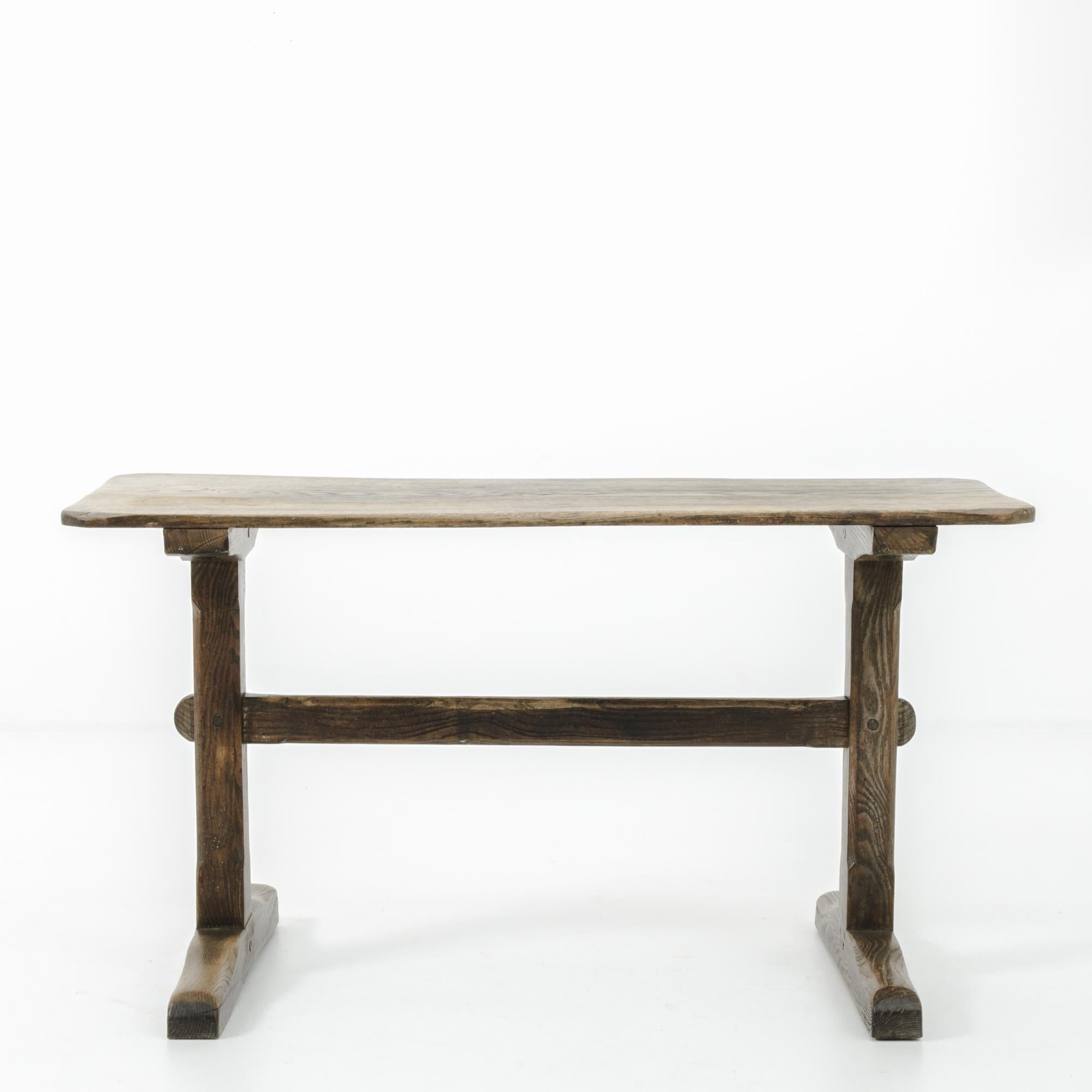 A rustic oak table from France, circa 1900. The silhouette is suggestive of a bistro or beer garden — a rough-hewn rectangular tabletop sits atop narrow trestle legs; wide feet provide a charming counterbalance. The wood is a dark brunette color,