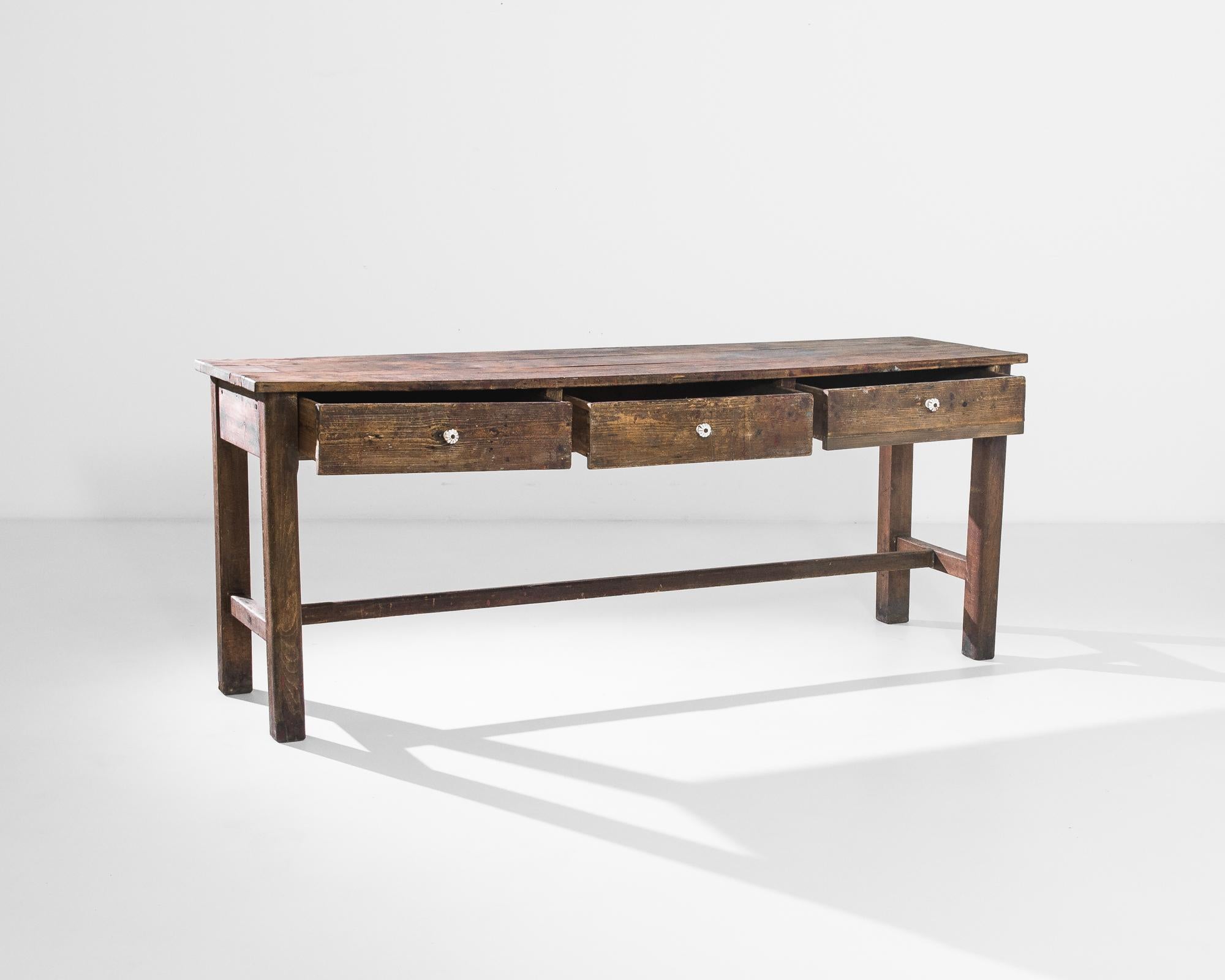 Elevate your living space with the charm of this 1900s French wooden console table, a harmonious blend of practicality and classic design. Crafted from wood, this table features three drawers along the side, each adorned with a sleek white knob for