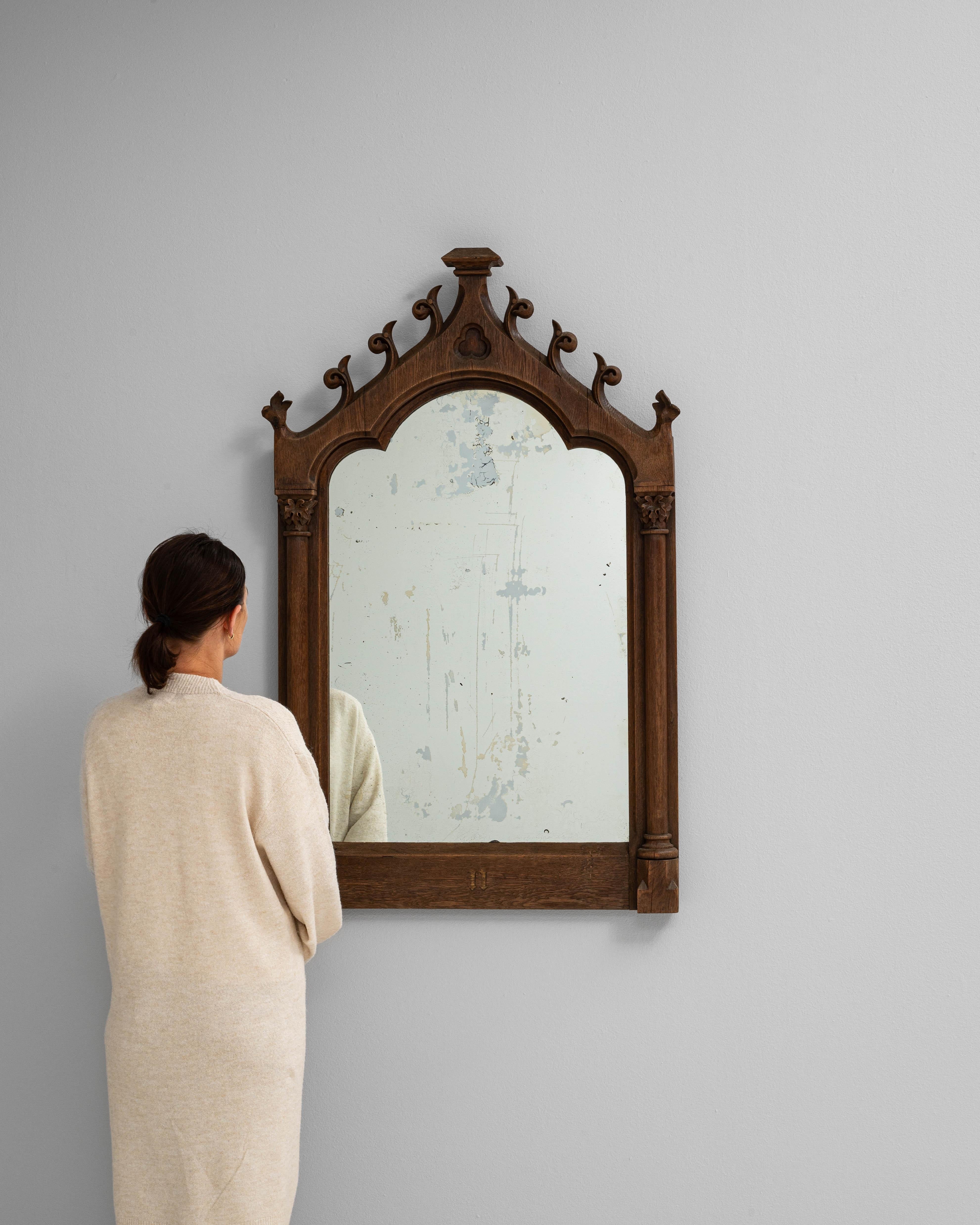 Ce miroir en bois français des années 1900 est un exemple frappant du style néo-gothique, avec un cadre arqué surmonté d'un fronton orné de volutes percées et de sculptures trilobées. Fabriqué en chêne richement grainé, le cadre est orné de colonnes