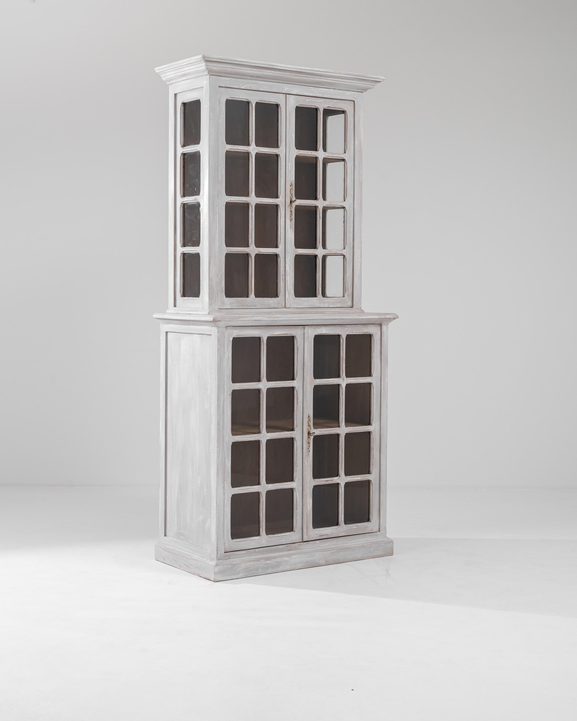 This wooden patinated vitrine was produced in France during the turn of the century. Featuring a double-case with glass doors, this tall vitrine features cornice beveled crown moldings, gilded scrolled escutcheons and a pearl gray distressed finish