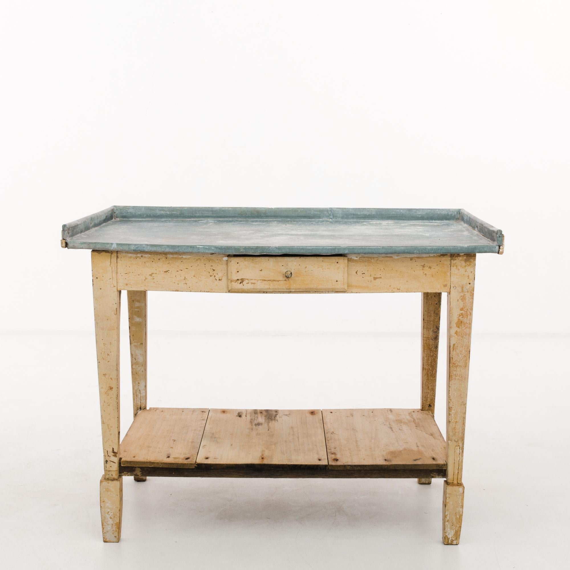 A wood patinated table with zinc top, produced circa 1900. A vintage work table standing on legs like four obelisks, featuring a sliding shelf under the tabletop, and a lower shelf of wood planks. The naturally anti-microbial and non-porous