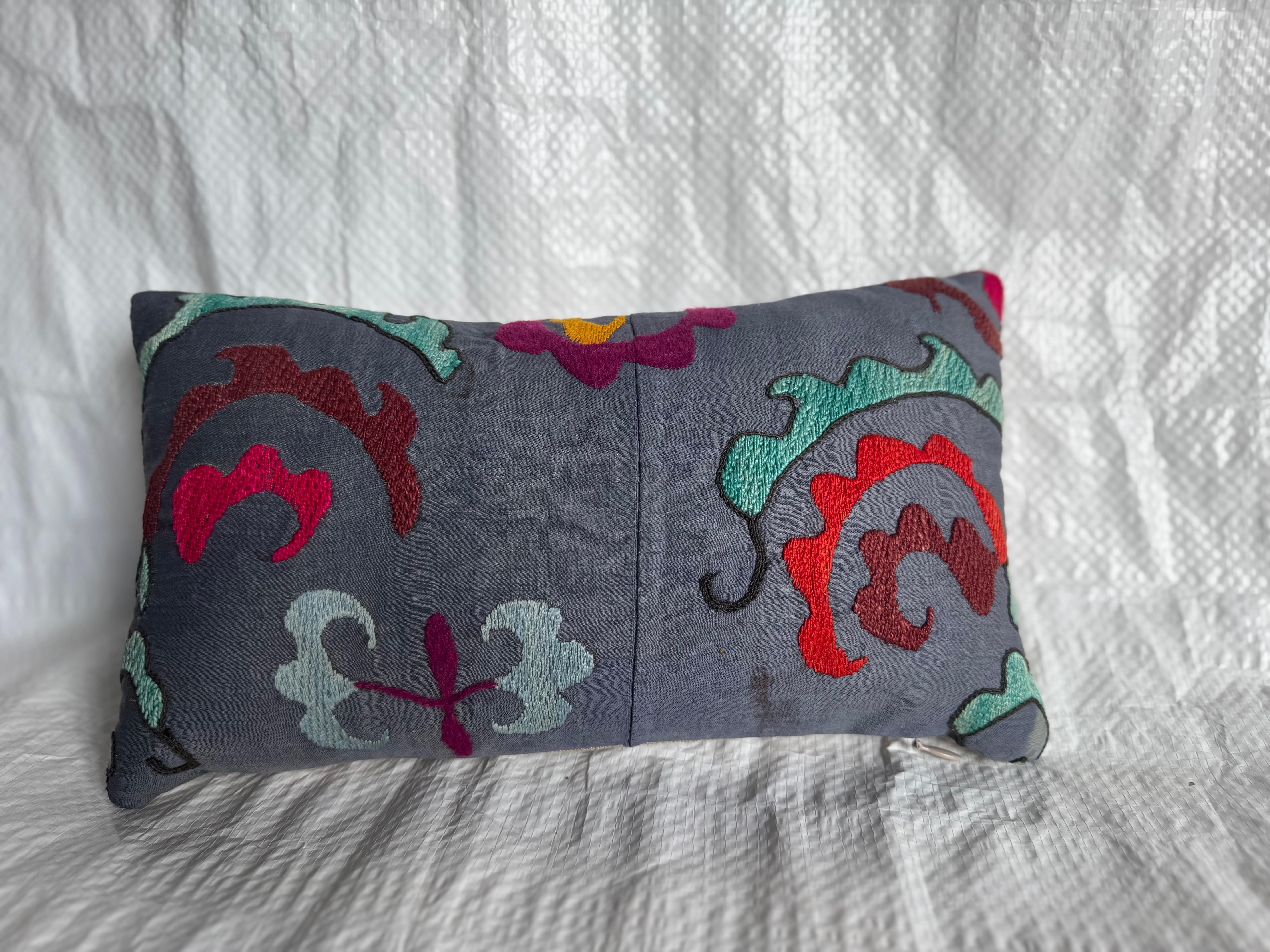 An exquisite early 1900s Suzani pillow featuring bold hand-embroidered floral and vine motifs in vibrant red, teal, ivory, and mustard hues on a soft charcoal-gray ground. This antique textile was originally part of a larger Central Asian Suzani — a
