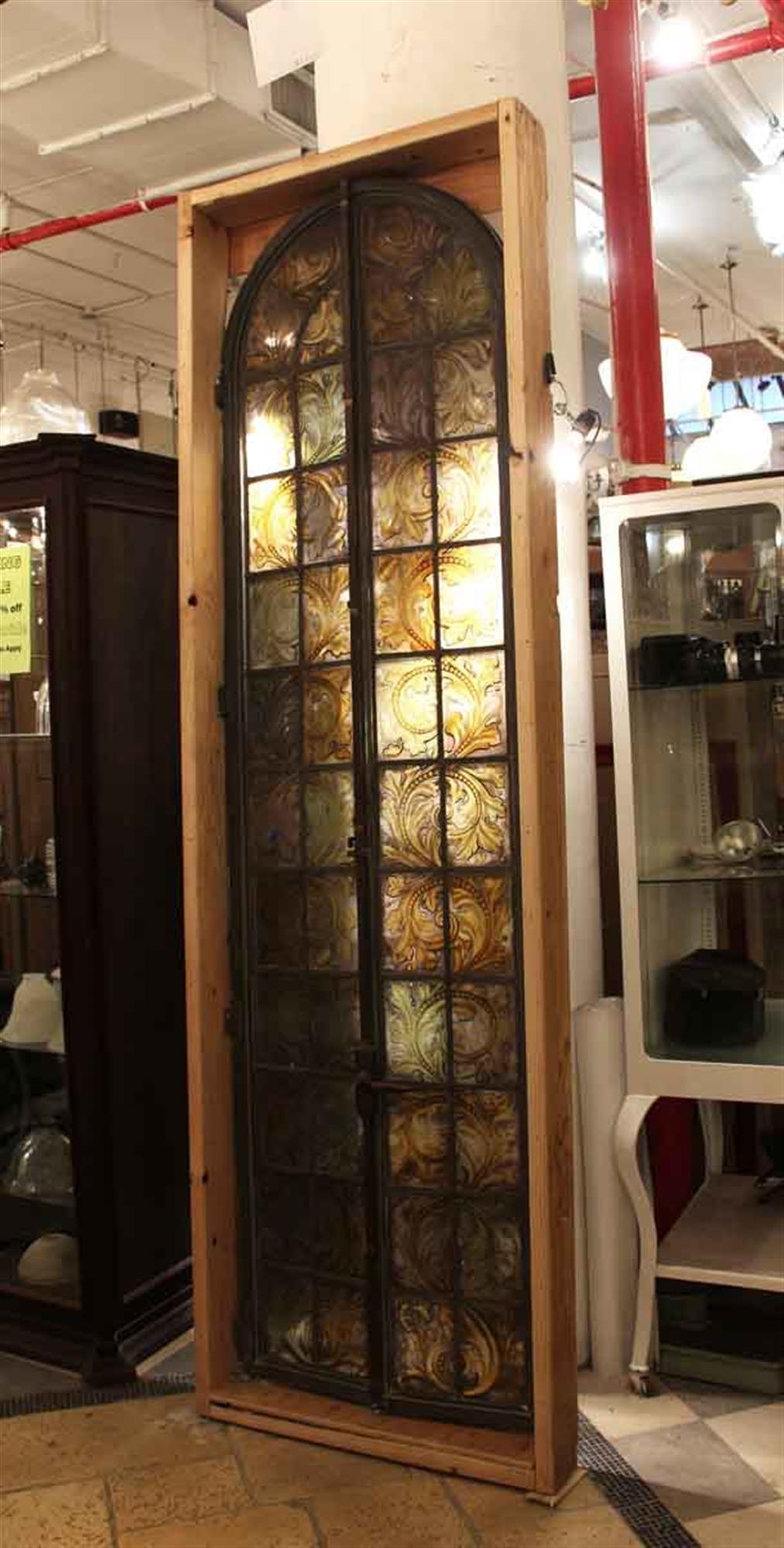 Stained glass arched bronze framed casement window. Secured in a wooden frame. There is a crack in the bottom left portion of the glass. Hand painted floral and leafy detail in amber tones. The dimensions are 98 in. H x 28 in. W, frame 102 in. H x