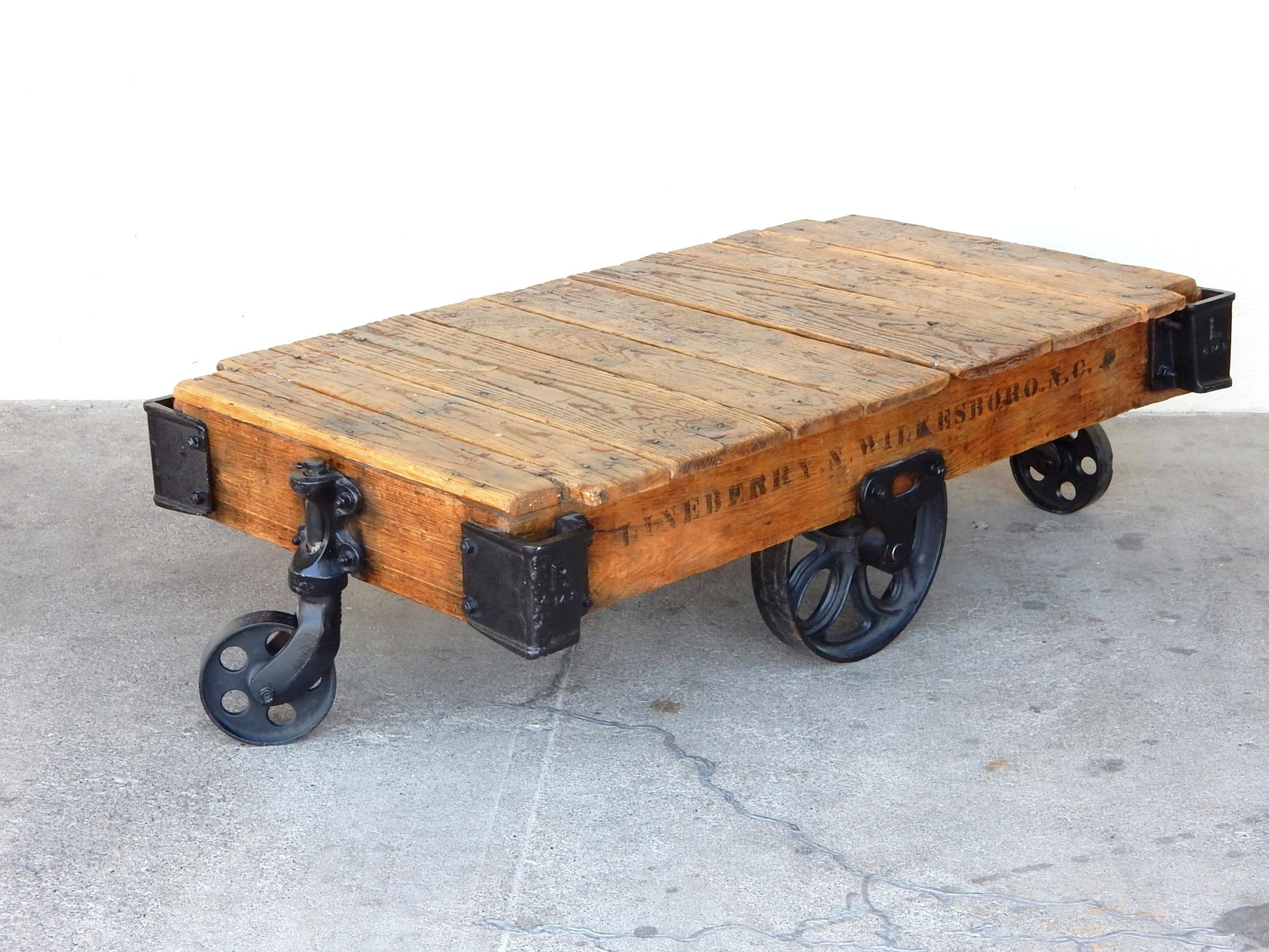 1900s Industrial oak and iron trolley cart refurbished to be a home coffee table.
Large central wheels with front and rear pivoting wheels. Sits at a slight angle. 
Stencil graphics on each side.
Solid heavy piece made of nailed oak planks on an