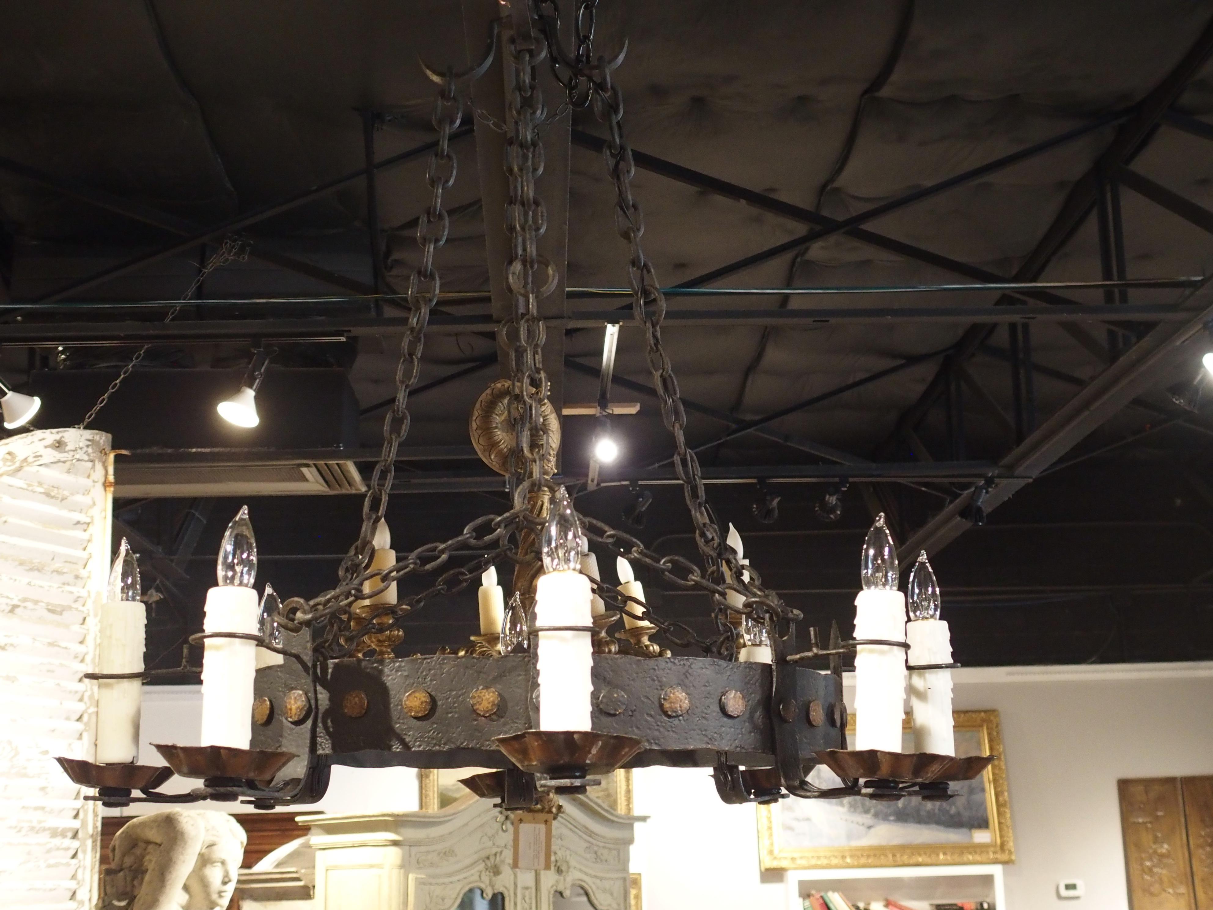 This antique French handwrought iron chandelier has a thick, hand-hammered circular band of ornamented metal with eight candleholders attached to the outside circumference. The bobeches are scalloped and have circular candle stabilizer wires about