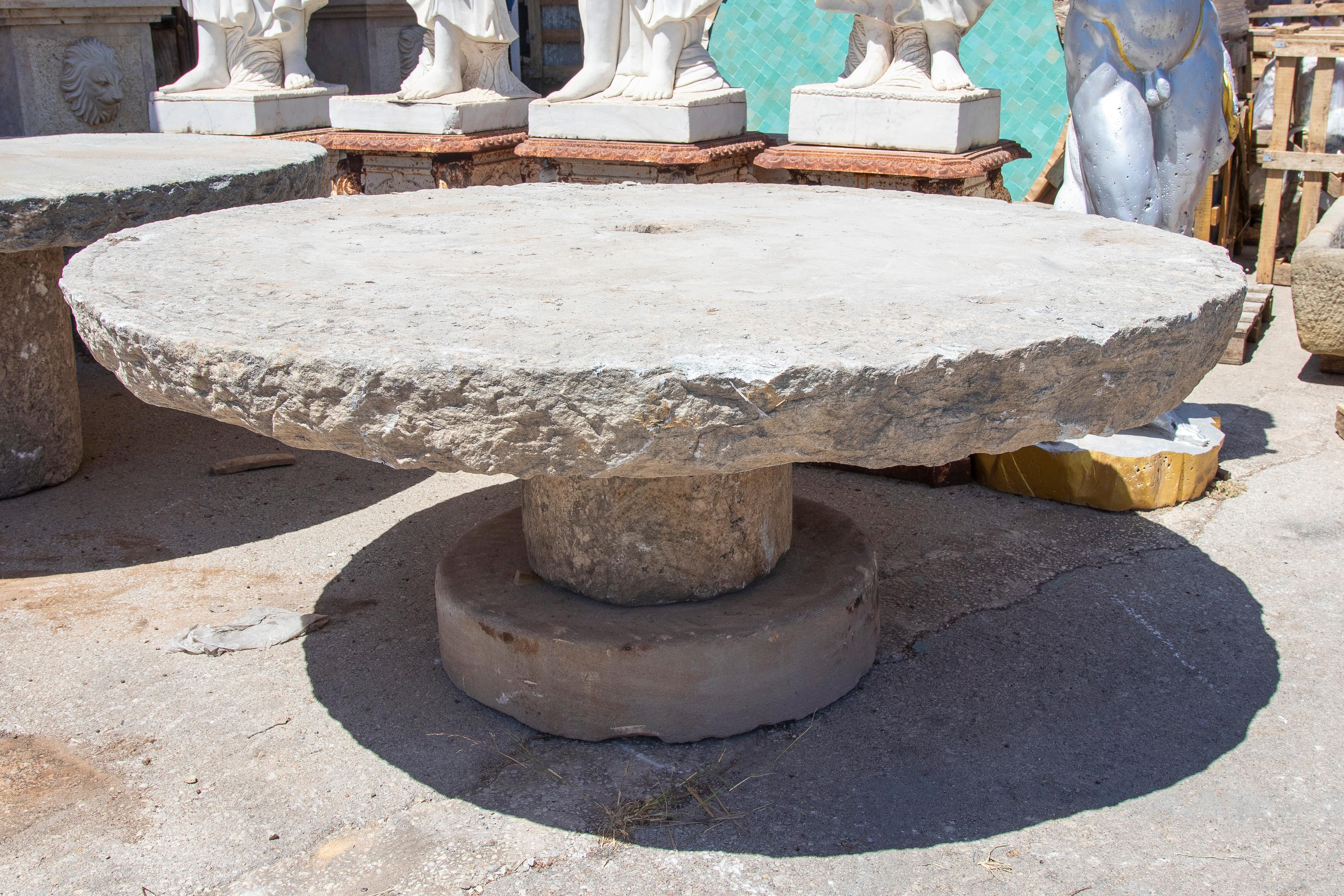 Esta imponente mesa de piedra natural es una pieza única que destaca por su estética rústica y su robusta construcción. Tallada a mano a partir de una sola pieza de piedra, presenta una superficie gruesa y circular con un borde irregular, que