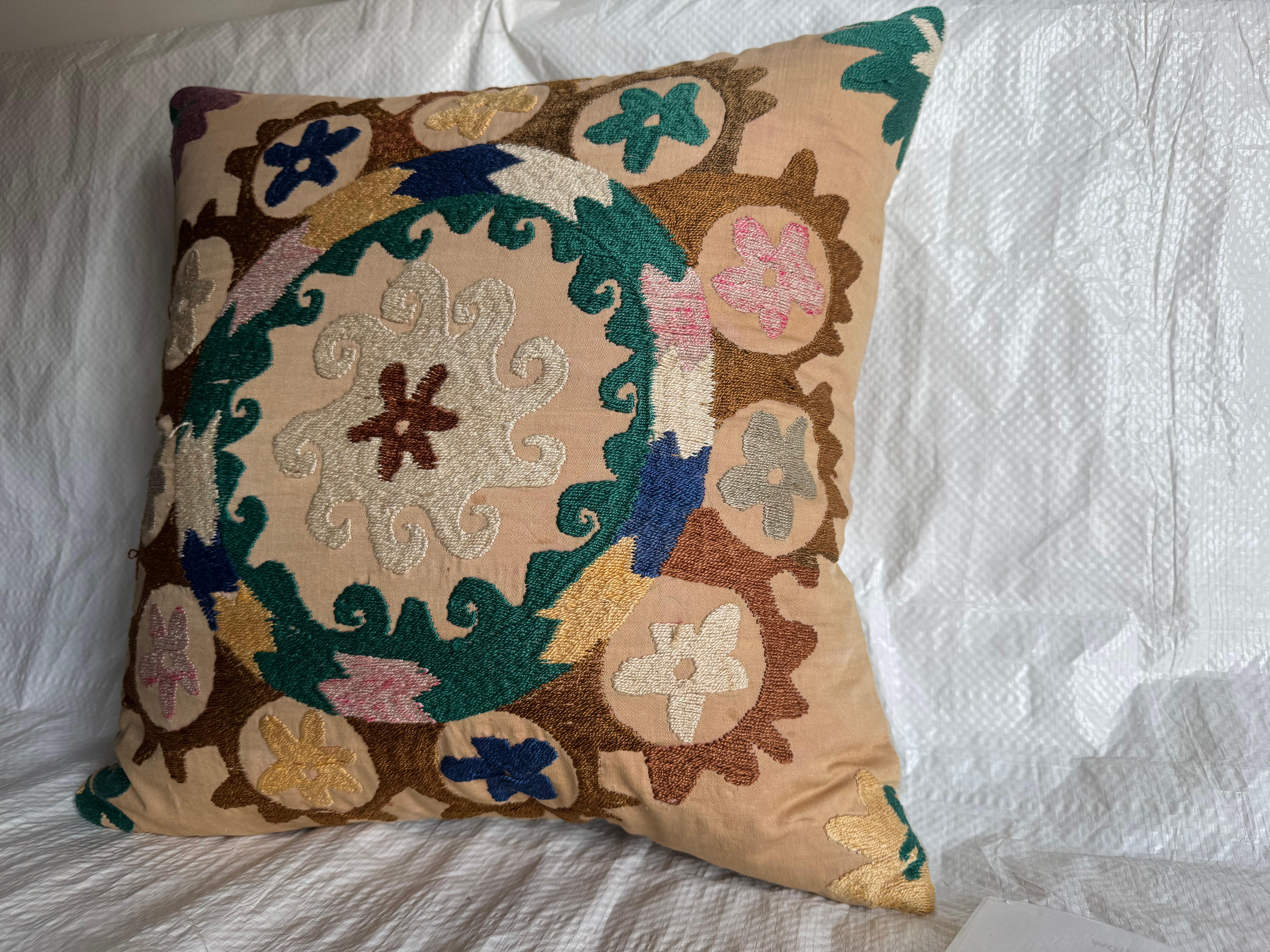 Cet oreiller unique est fabriqué à partir d'un textile Suzani brodé à la main et datant du début du XXe siècle. Le motif est centré sur un médaillon ivoire tourbillonnant entouré de motifs floraux et de vignes dans les tons sarcelle, or, marron et