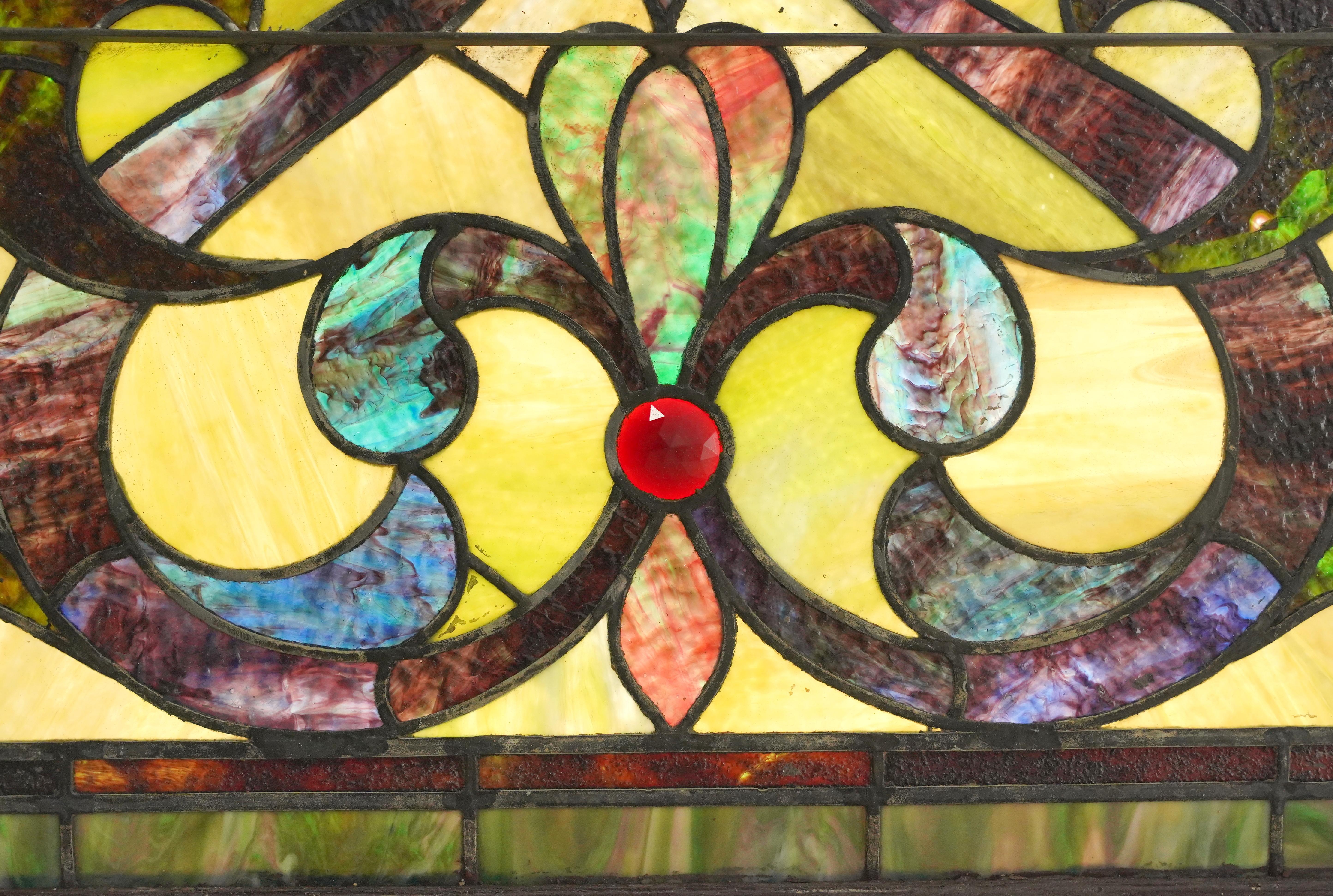 Antique arched church window with textured stained glass panels in multi colored swirl arrangements with a red jewel center. The outside frame is painted a reddish-orange color, better known as terra cotta. It was acquired from a church in