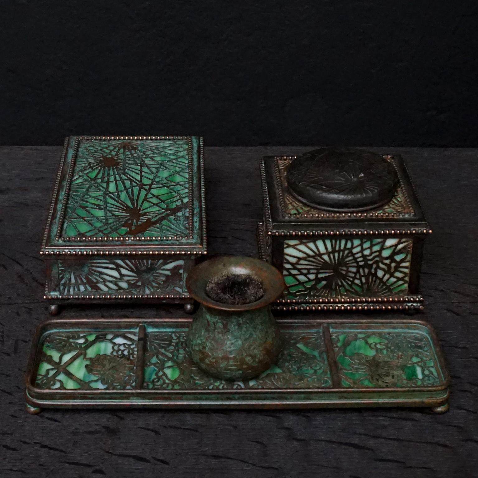Green mottled slag glass and etched bronze desk set by Tiffany Studios New York, consisting of a little hinged lid box a lidded inkwell both in the Pine needle pattern and a pen wipe and pen tray both in the Grapevine pattern. 
Each piece stamped