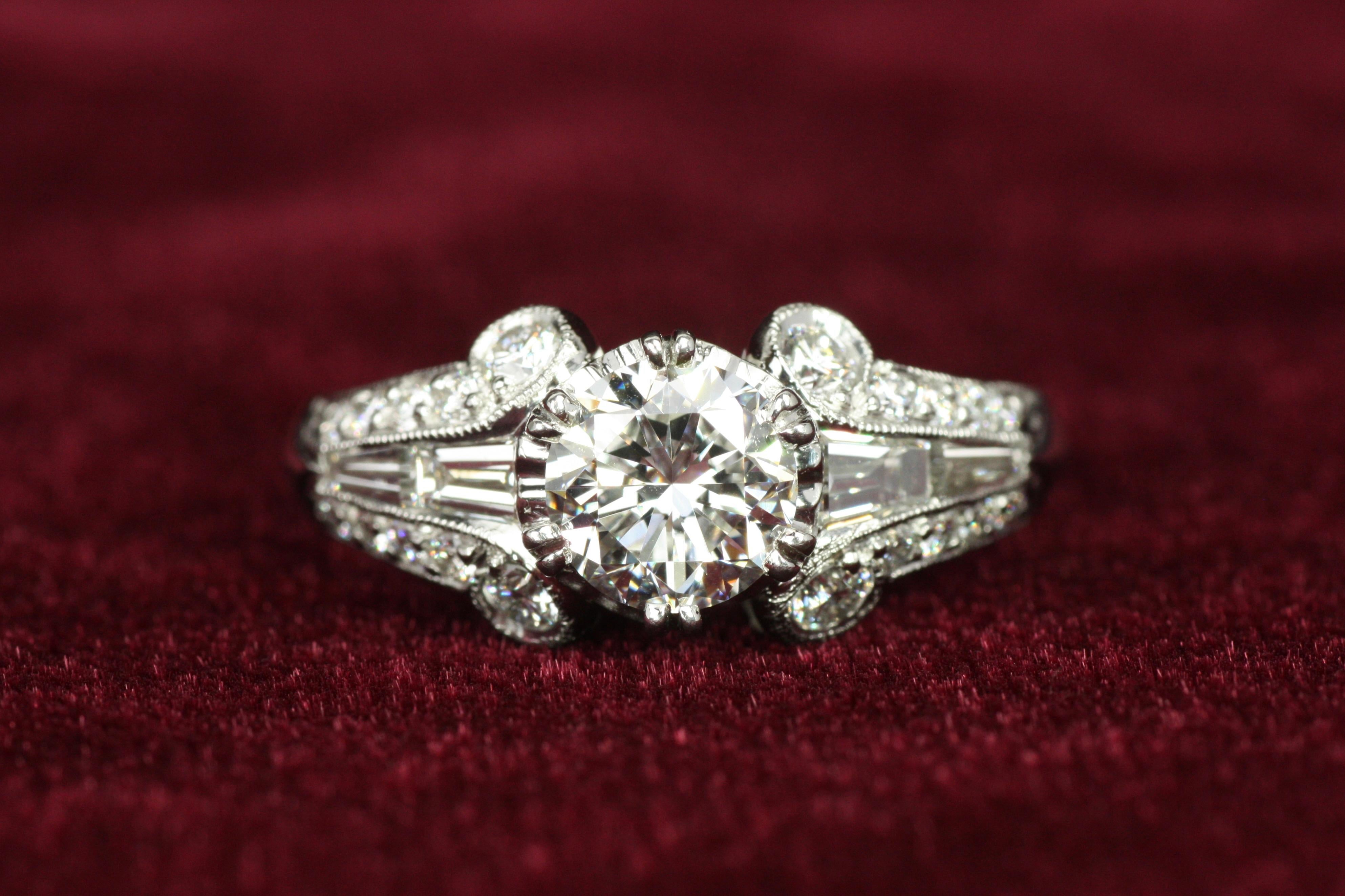 This stunning Art Deco engagement ring dazzles with timeless elegance, showcasing a 1.05ct round brilliant cut diamond as its centerpiece. Its geometric baguette and round cut diamond accents, knife-edge band, and milgrain details lend a bold yet