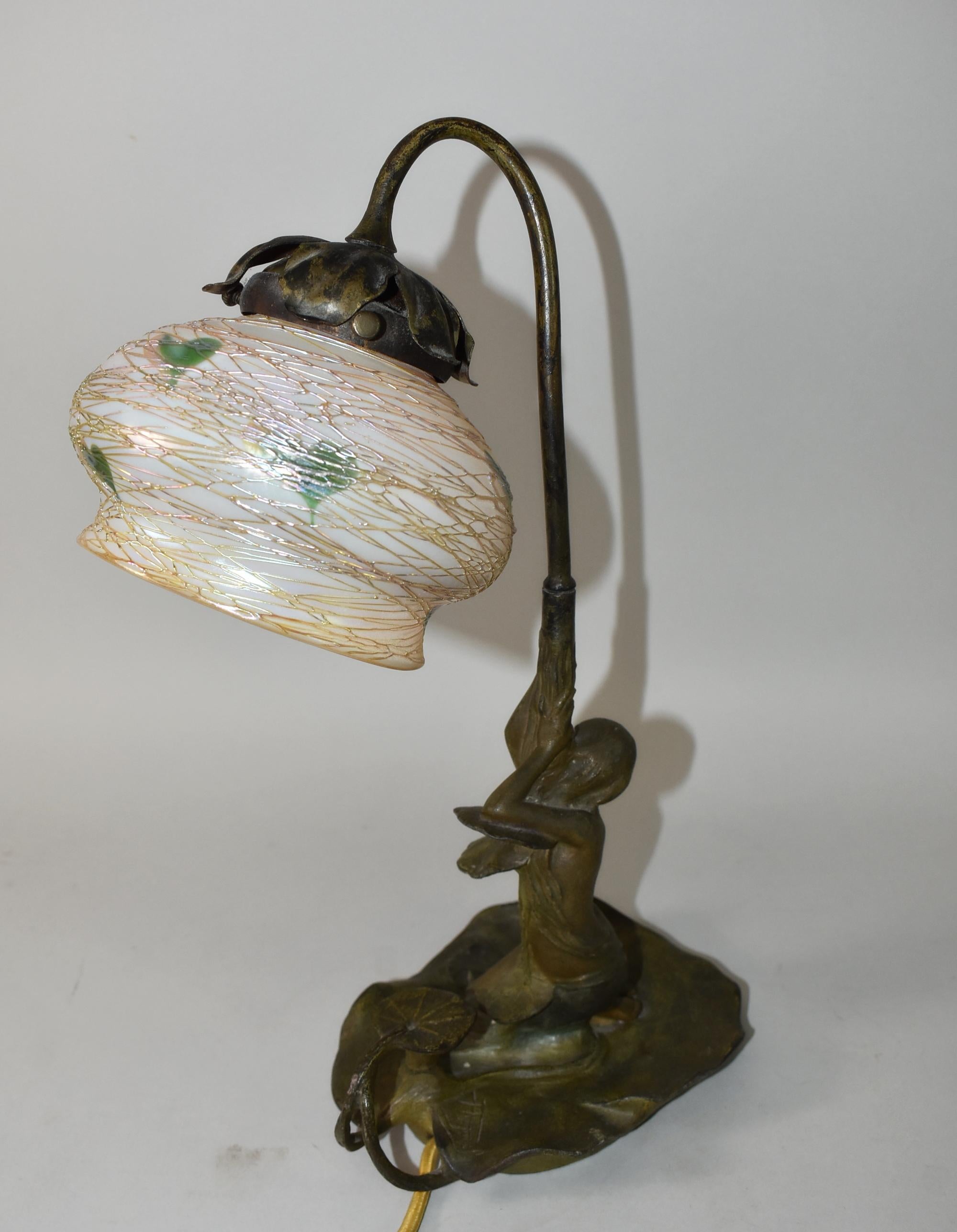 This turn of the century art nouveau boudoir lamp by P. Rigual is fitted with a Quezal shade. The bell form iridescent glass shade has gold threaded heart and vine decoration on a figural patinated spelter base, depicting a nymph on a lily pad. The