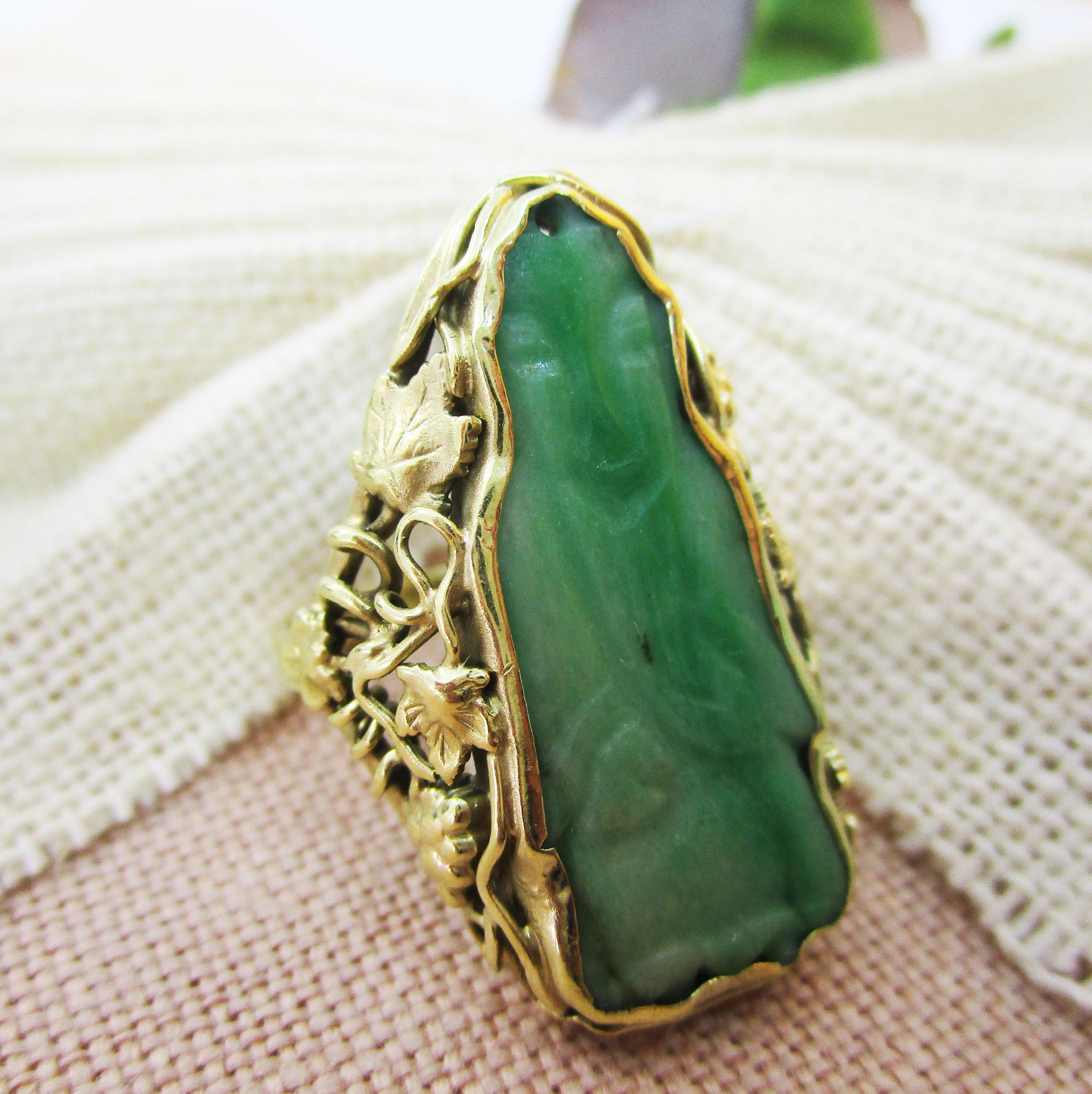 This killer Arts and Crafts ring is in 14k green gold and features a stunning carved jade center! The ring has a distinctly Arts and Crafts design that is clear in the curling vines of the grapevine and leaf motif that makes up the shoulders and