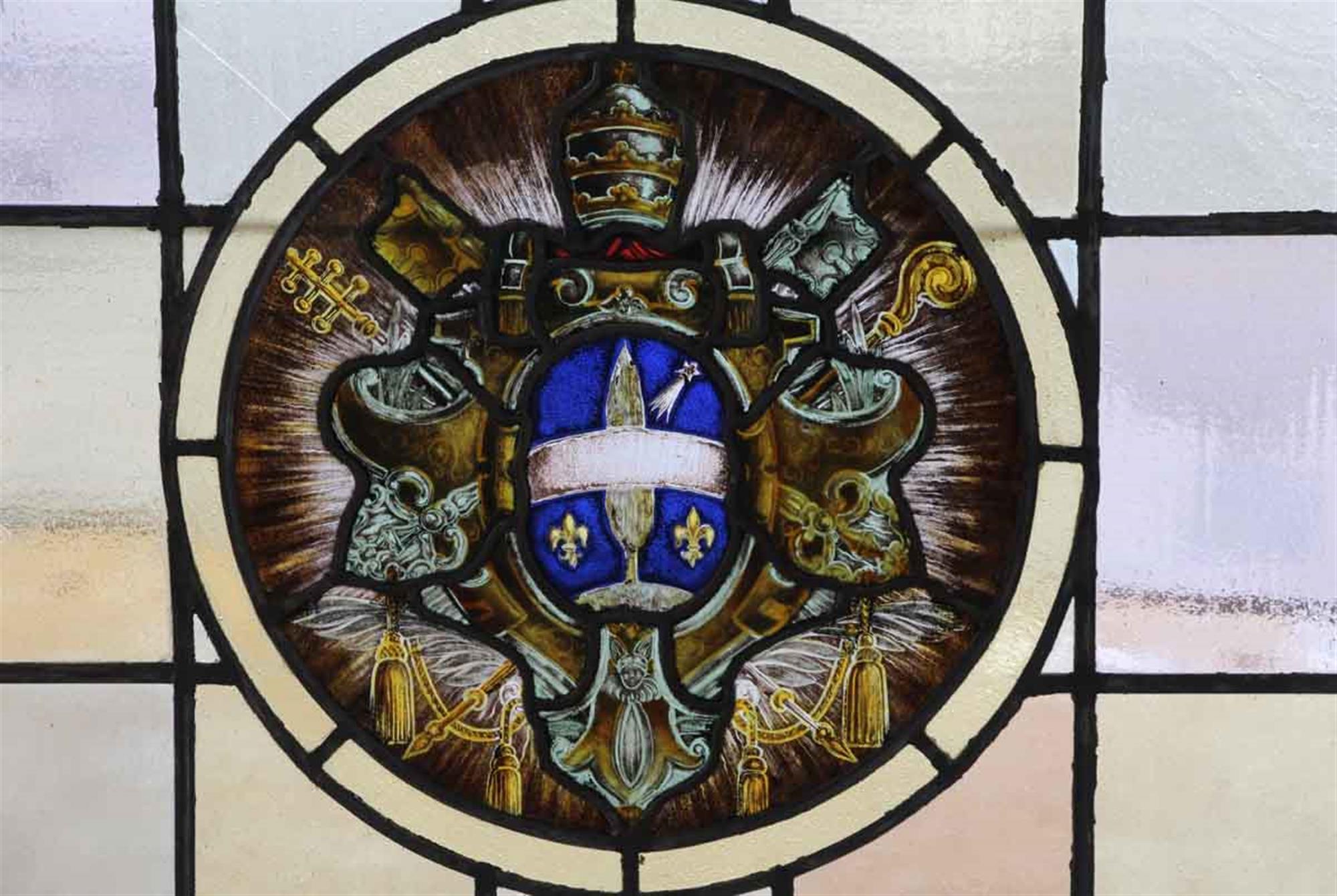 Wood framed 1910 Arts & Crafts stained glass window with a center painted shield, fleur-de-lis and shooting star motif detail. One pane of glass just outside the circle has a stress crack. This can be seen at our 302 Bowery location in Manhattan.