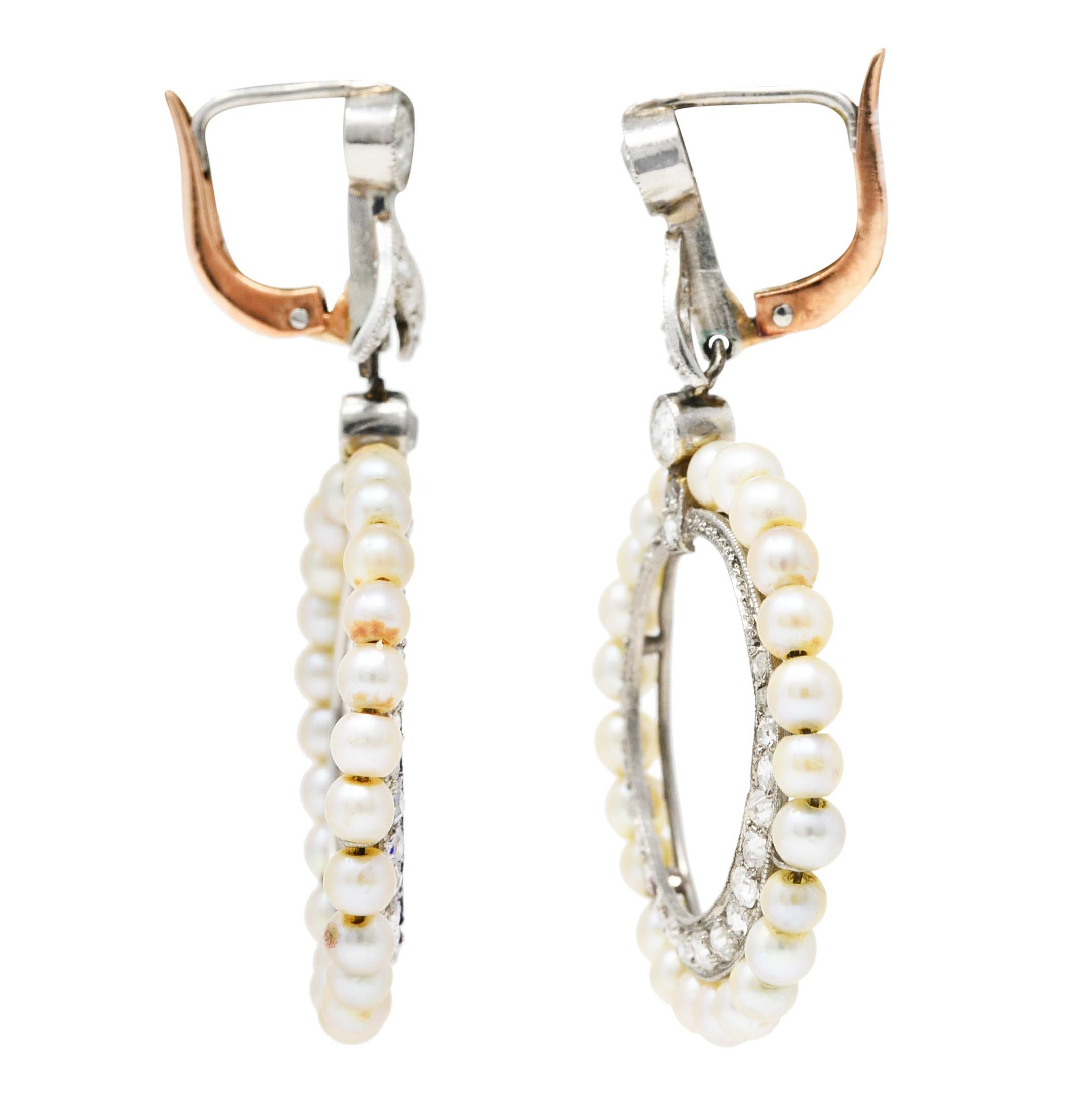 Earrings are designed as diamond foliate surmounts suspending an articulated drop. Drop is two concentric circles with an inner diamond halo surrounded by a halo of strung 3.0 mm pearls. Pearls are well matched in cream body color with good to very