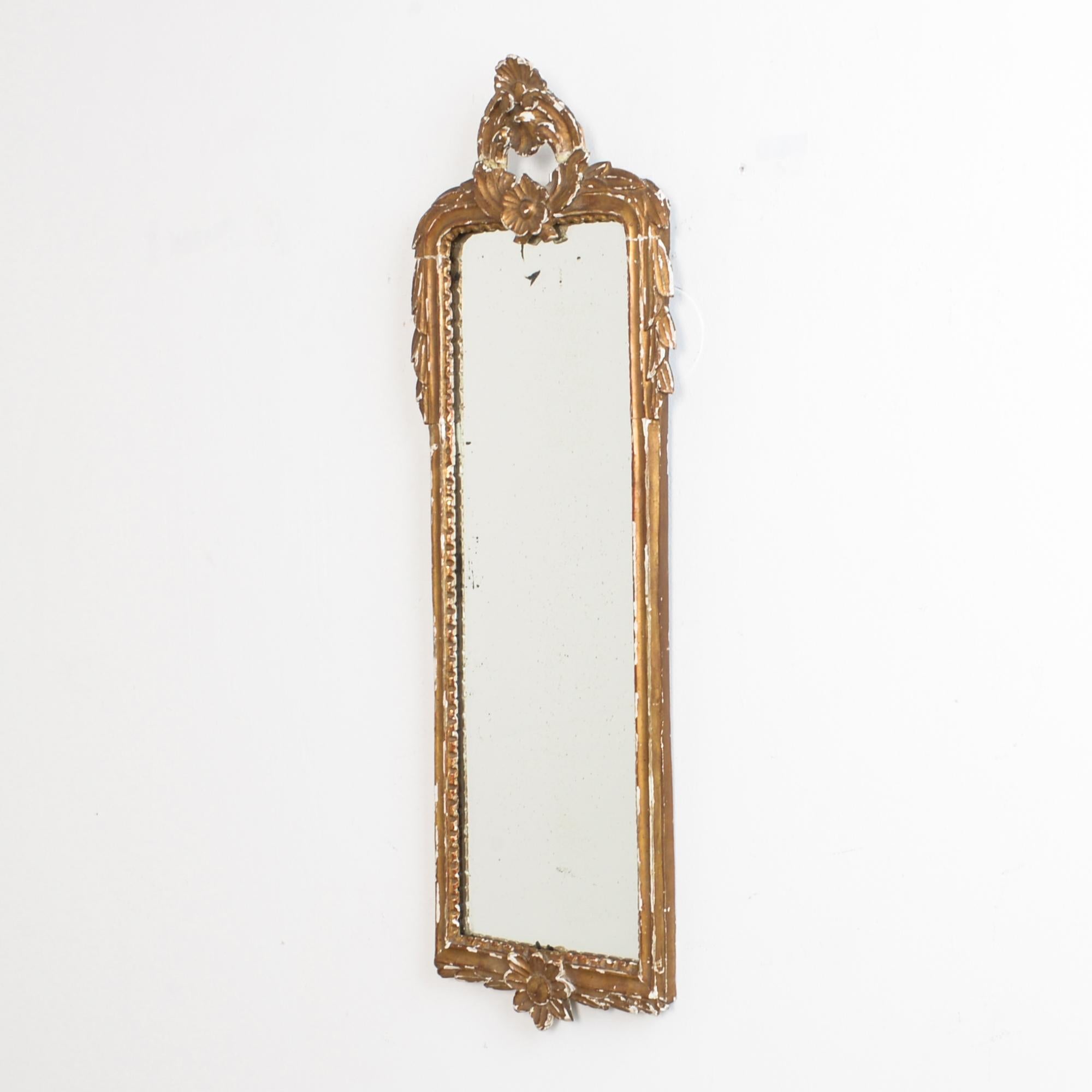 From France, circa 1910. A worn patina reveals the layers, a striking white plaster ground. Carved in an architectural style, floral elements accentuate the slender shape of the mirror. Affixed above and below the mirror is further framed with