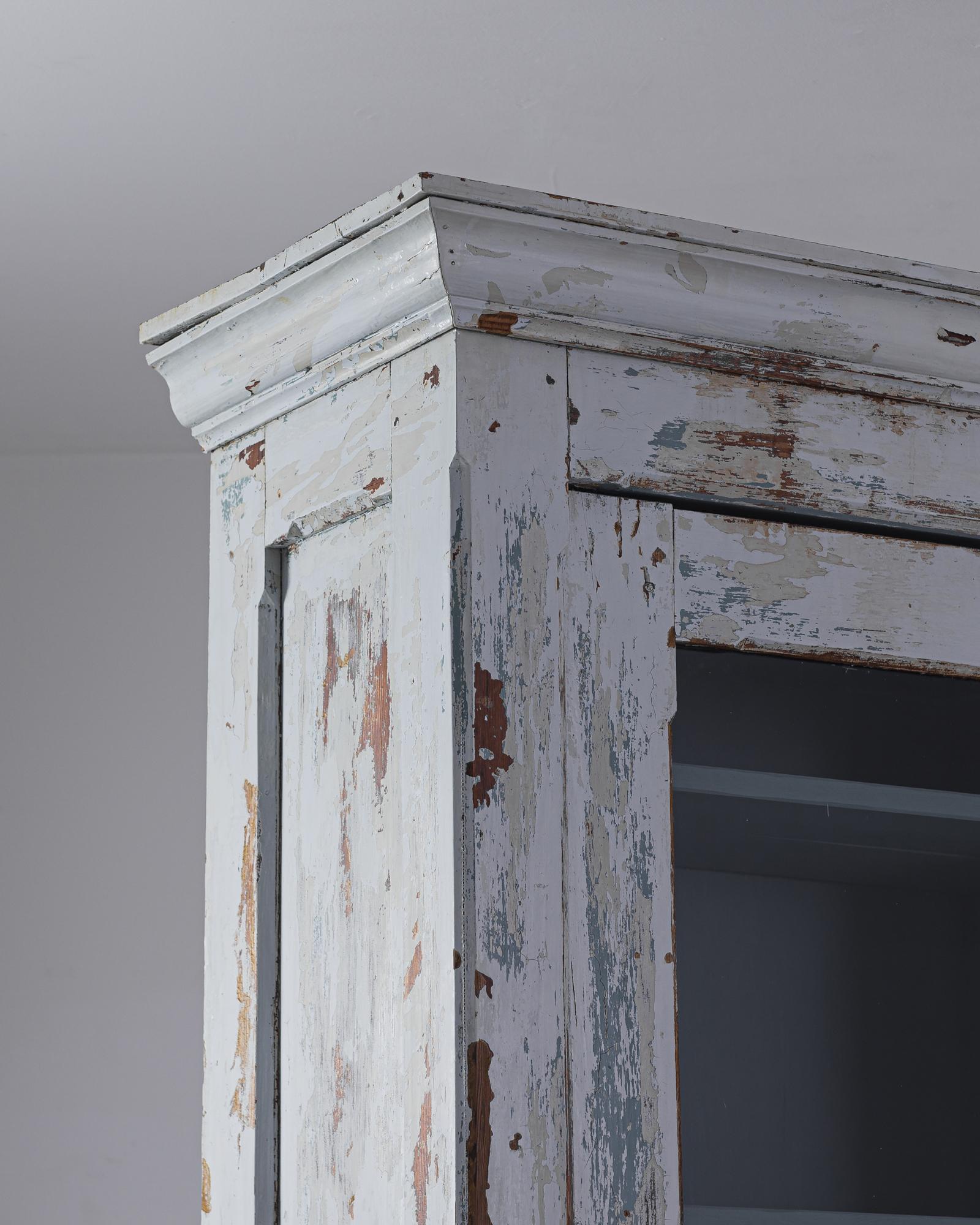 A wooden vitrine from 1910s Belgium with an evocative patina. A tall, slender cabinet, fronted by a paned glass door, sits atop a low cupboard. The original periwinkle paint has weathered to reveal notes of pigeon-grey, teal, and natural wood; a