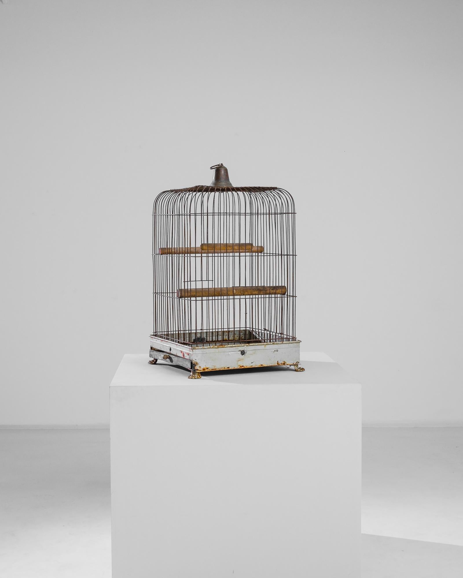 A metal birdcage from France, produced circa 1910. An antique birdcage standing two feet eight inches, a round edge rectangle of rusting bars rising to bell shaped cupola, wrapped at its base by a white metal band resting on four clawed feet.