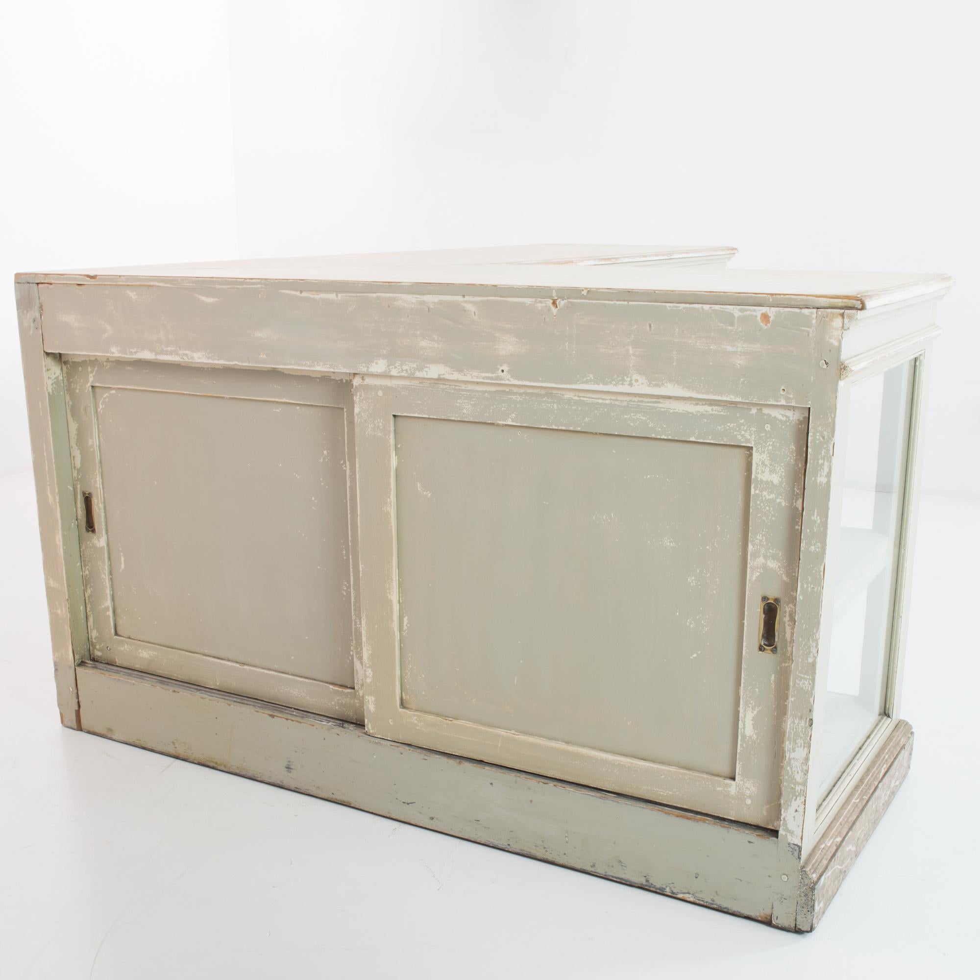 An L-shaped wooden shop counter from France, circa 1910, with a nostalgic patina. Glass windows on the front of the case allow a full view of the items on display; in the back, sliding doors provide access to the interior shelves. The front and