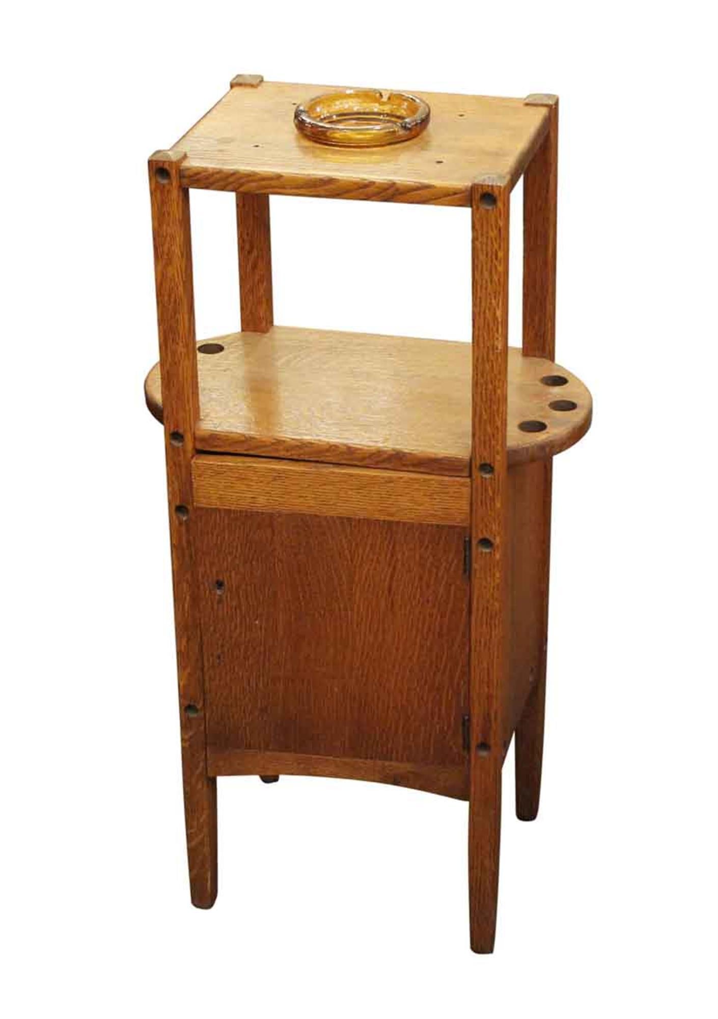 1915 small freestanding side smoking table with a combination cabinet and key. It is made of natural oakwood with an original finish and a simple Arts & Crafts design. Included is an amber glass ashtray and six holes for pipes. This can be seen at