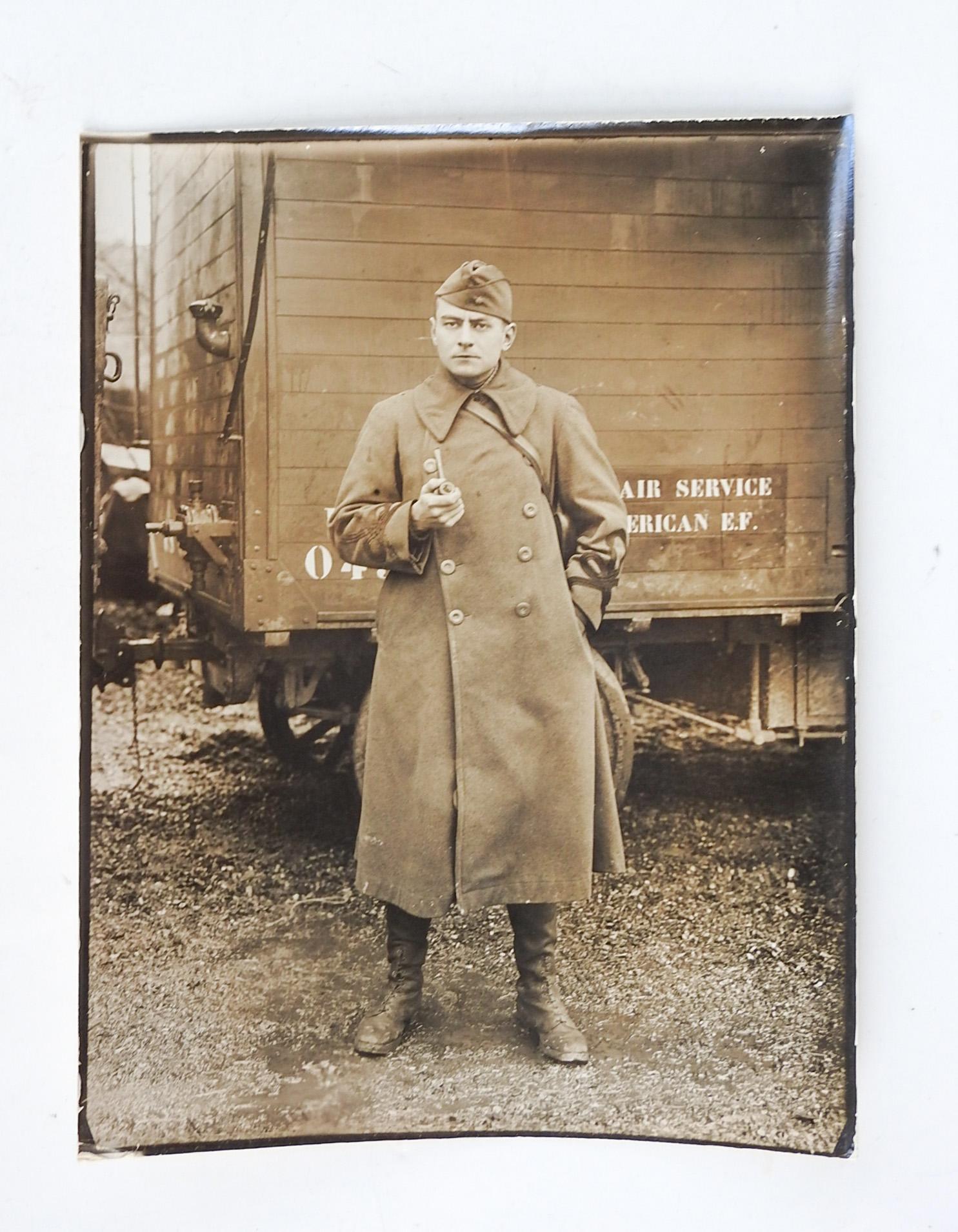 Circa 1918 silver gelatin photograph of US soldier or pilot. Standing in front of a transport for Air Service American Expeditionary Force. Lace up boots, heavy wool coat with braid at sleeves and cap. Holding a pipe and with a tough expression,