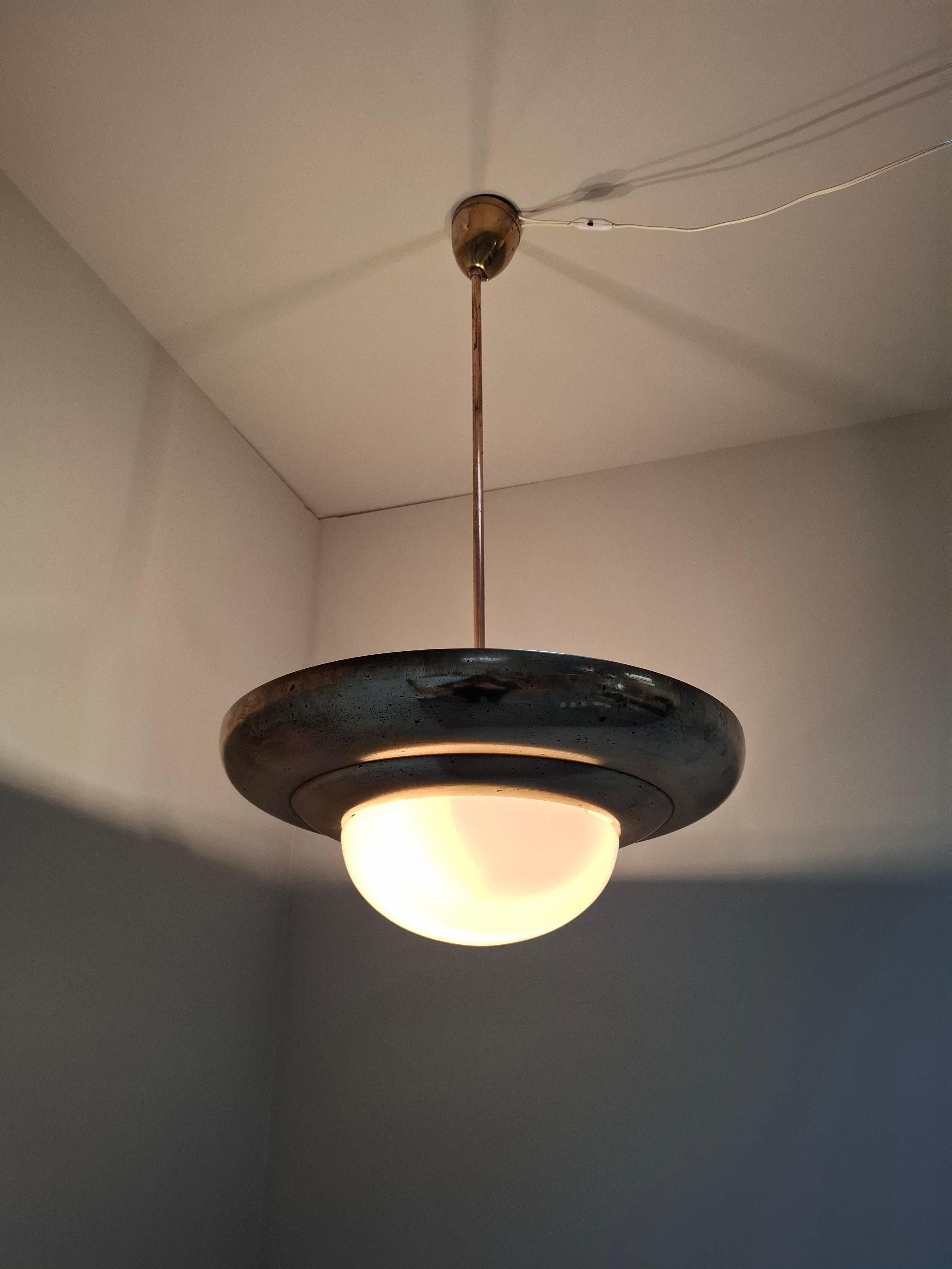 Gran lámpara colgante Art Decó diseñada por Napako, tipo 13443, fabricada en Checoslovaquia hacia 1920. La luminaria presenta un escultural cuerpo de latón con auténtica pátina envejecida y una llamativa cúpula de cristal de leche opalino que