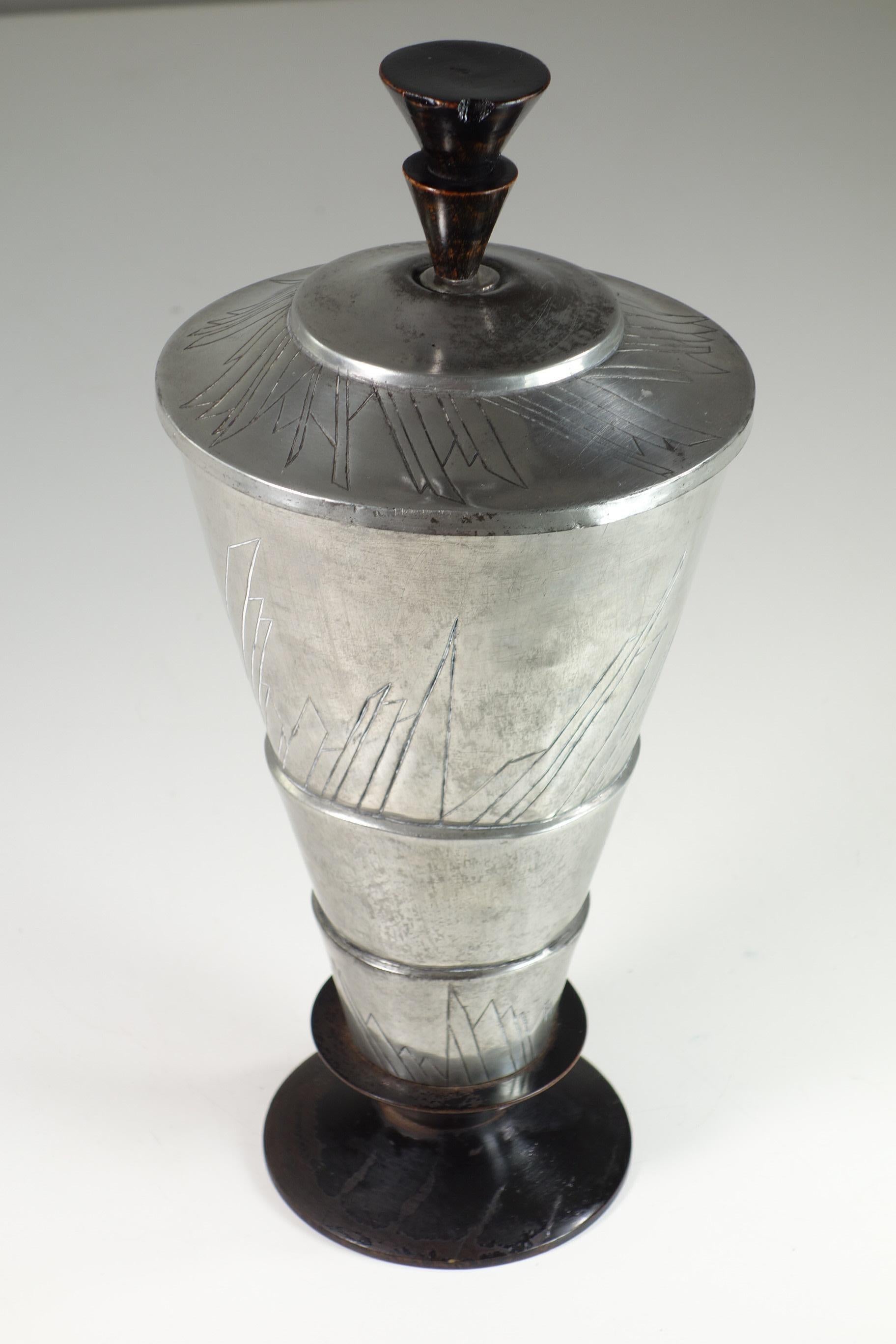 This is a Futurist urn from the 1920s, crafted from metal and black lacquered wood. Its bold, conical shape and dynamic geometric engravings reflect the influence of the Futurist movement, emphasizing speed, energy, and modernity. The contrast