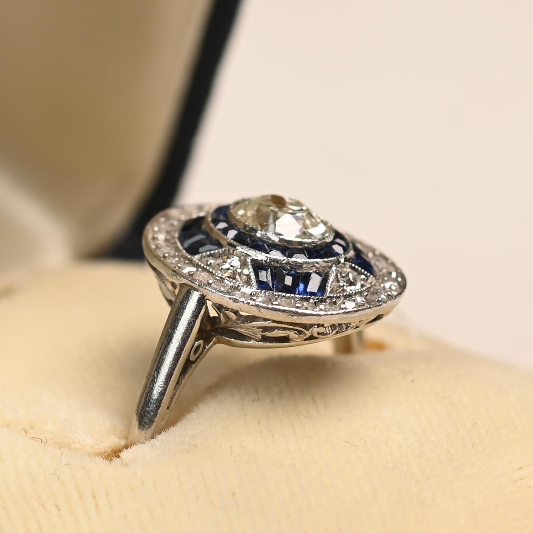 Dieser exquisite Ring aus der Art-Déco-Ära ist mit einem antiken Diamanten im Ovalschliff besetzt, der etwa 0,5 Karat wiegt. Der Diamant wird von Halos aus Saphiren im Kaliberschliff und funkelnden Diamanten akzentuiert, die in einem auffälligen