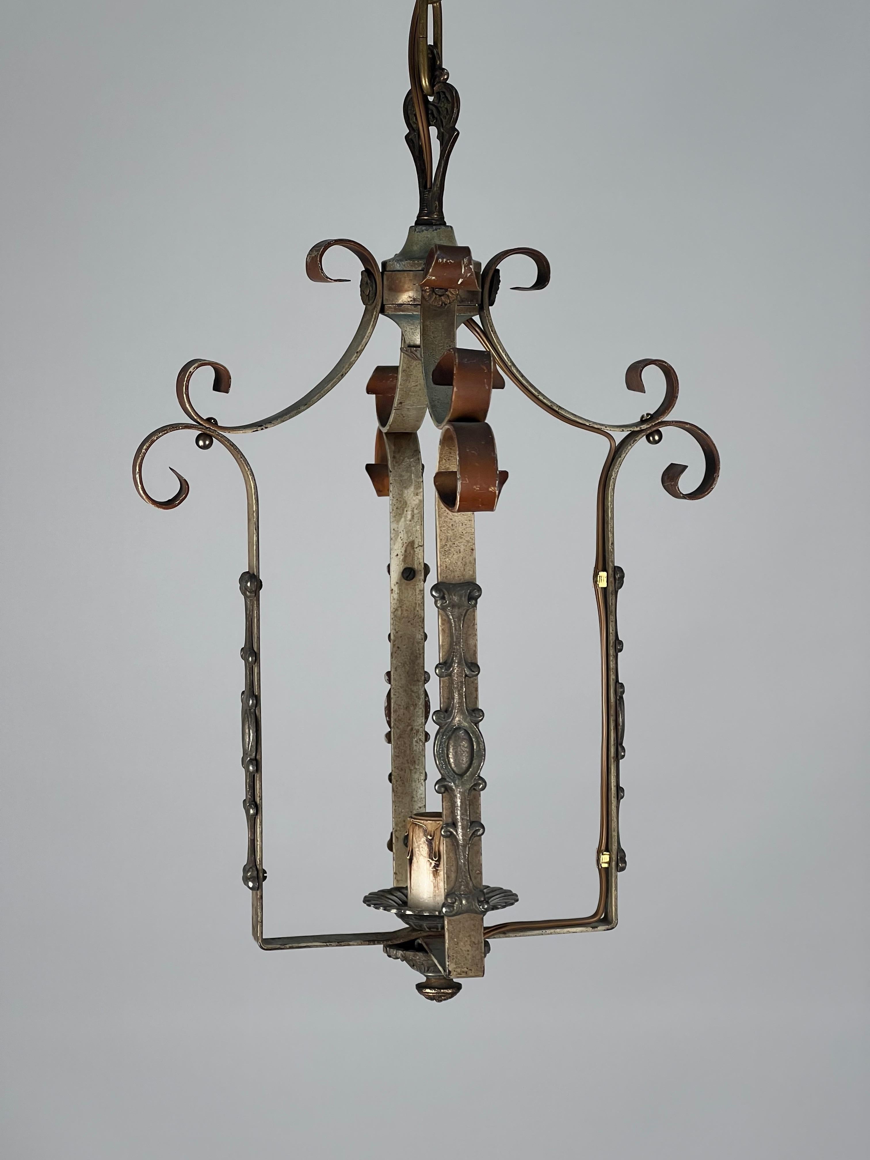 1920's American open lantern featuring suggestions of Tudor revival scrolls and shield appliques. 

A central single medium base socket illuminates each of the four strap arms. We have chosen to leave the finish and patina as is because of its